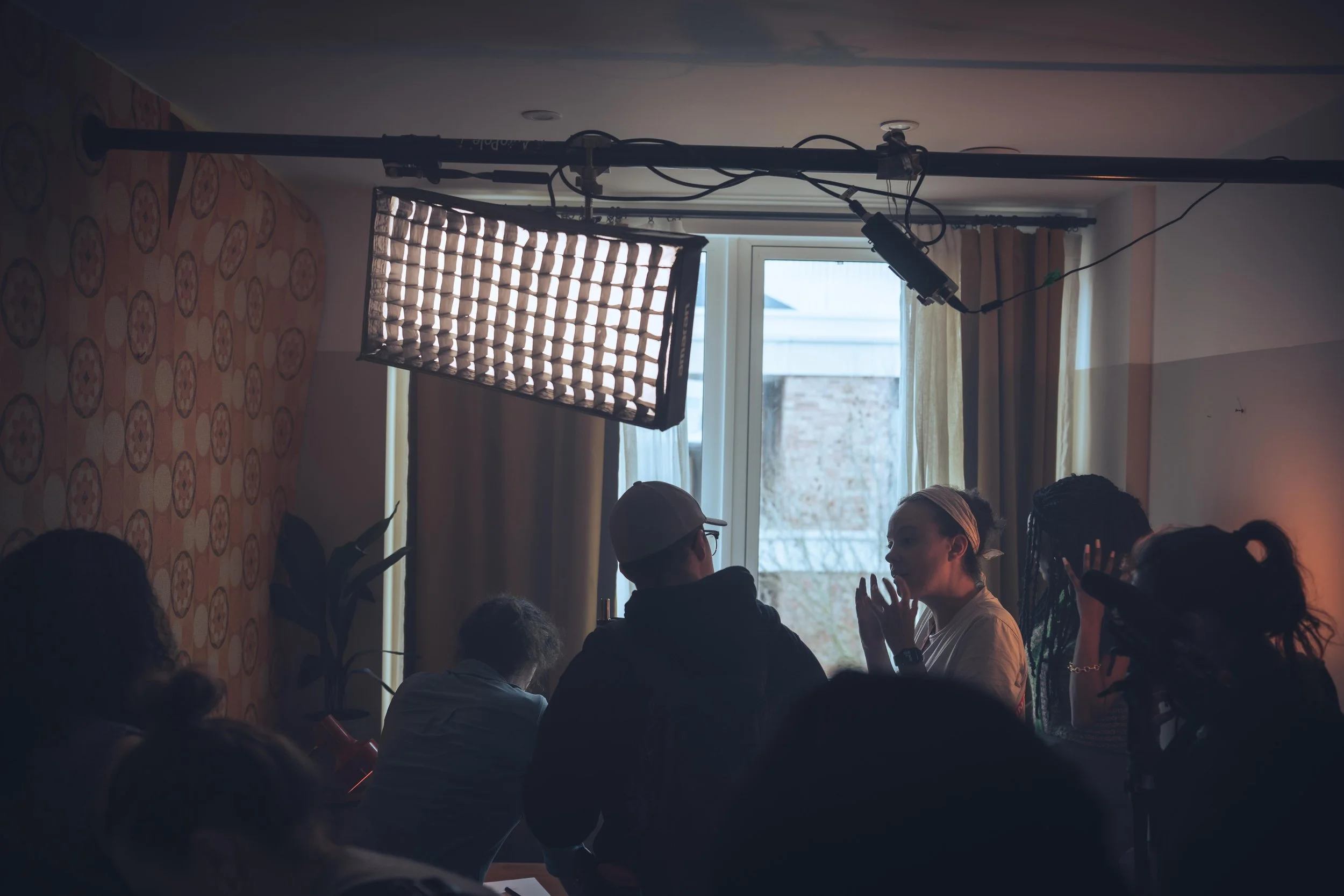 Group of people sitting and talking indoors near a window with curtains, with lighting equipment set up overhead.
