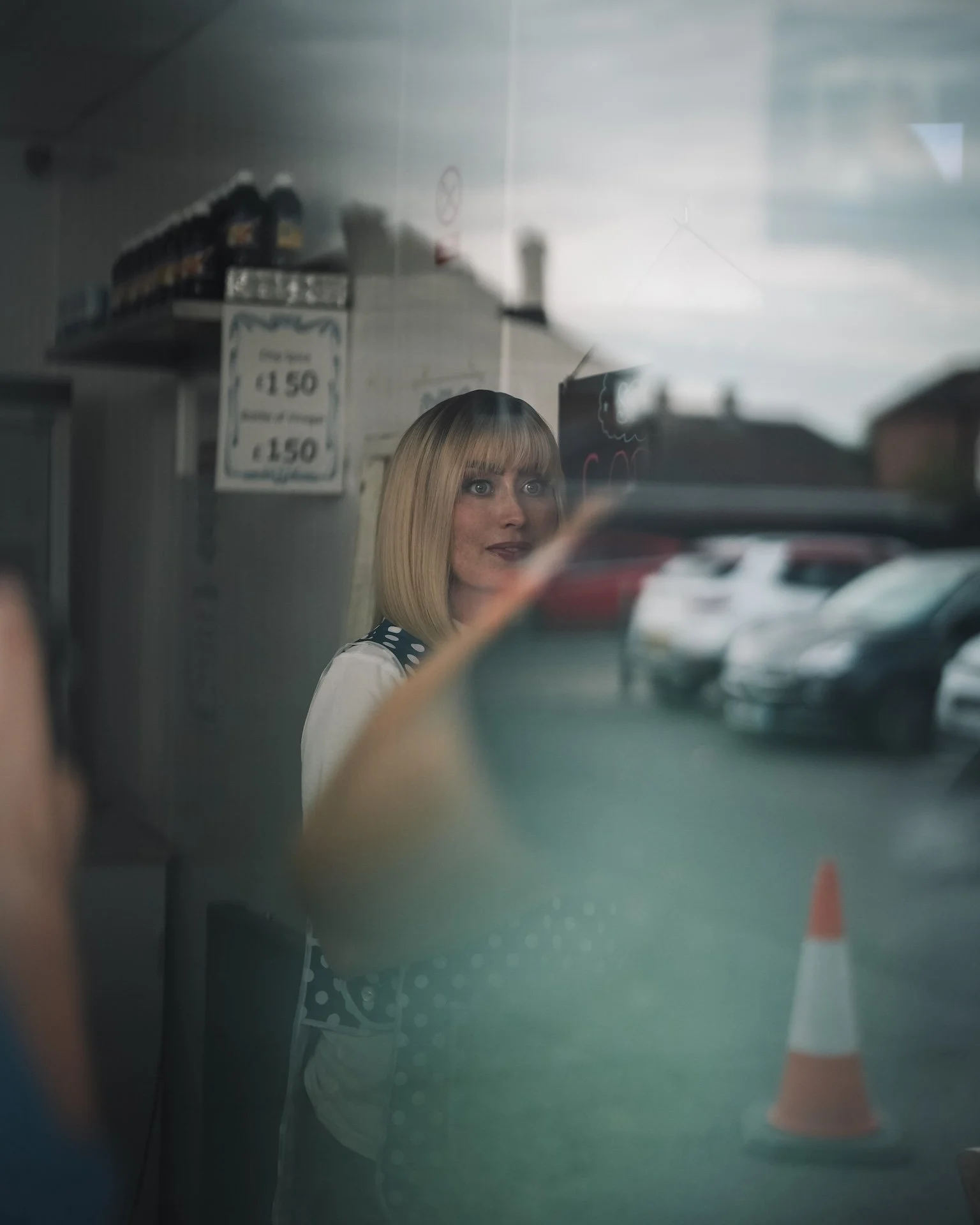 Reflection of a woman with blonde hair and bangs taking a photo in a store window with a traffic cone and cars outside.