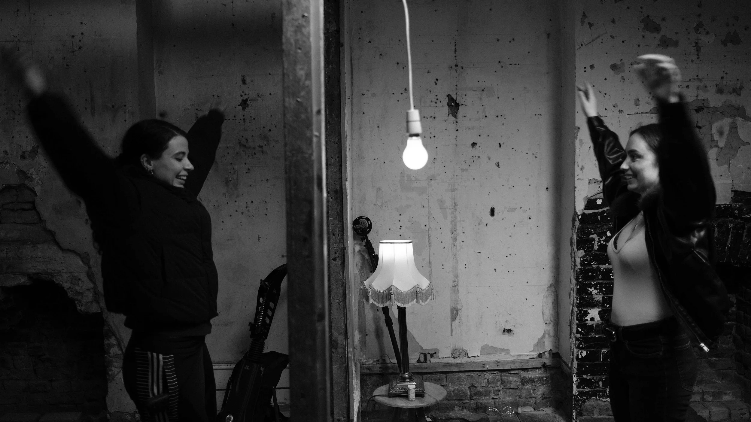 Two women smiling with arms raised, facing each other in a room with exposed brick and concrete walls, one with a jacket and the other with a leather jacket, illuminated by a hanging light bulb and a table lamp.
