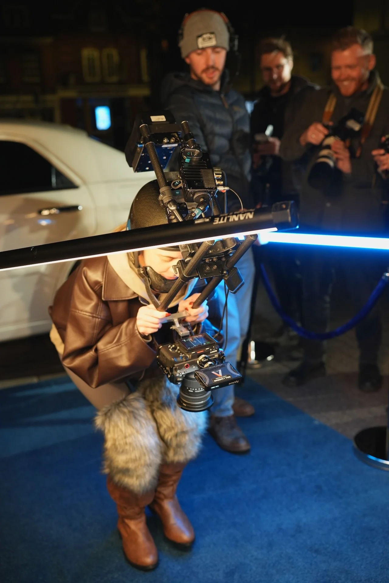 A person operating a professional camera mounted on a tripod in a dark indoor setting, with a child in brown boots and furry coat standing nearby and a group of three men in the background, some holding phones and smiling.