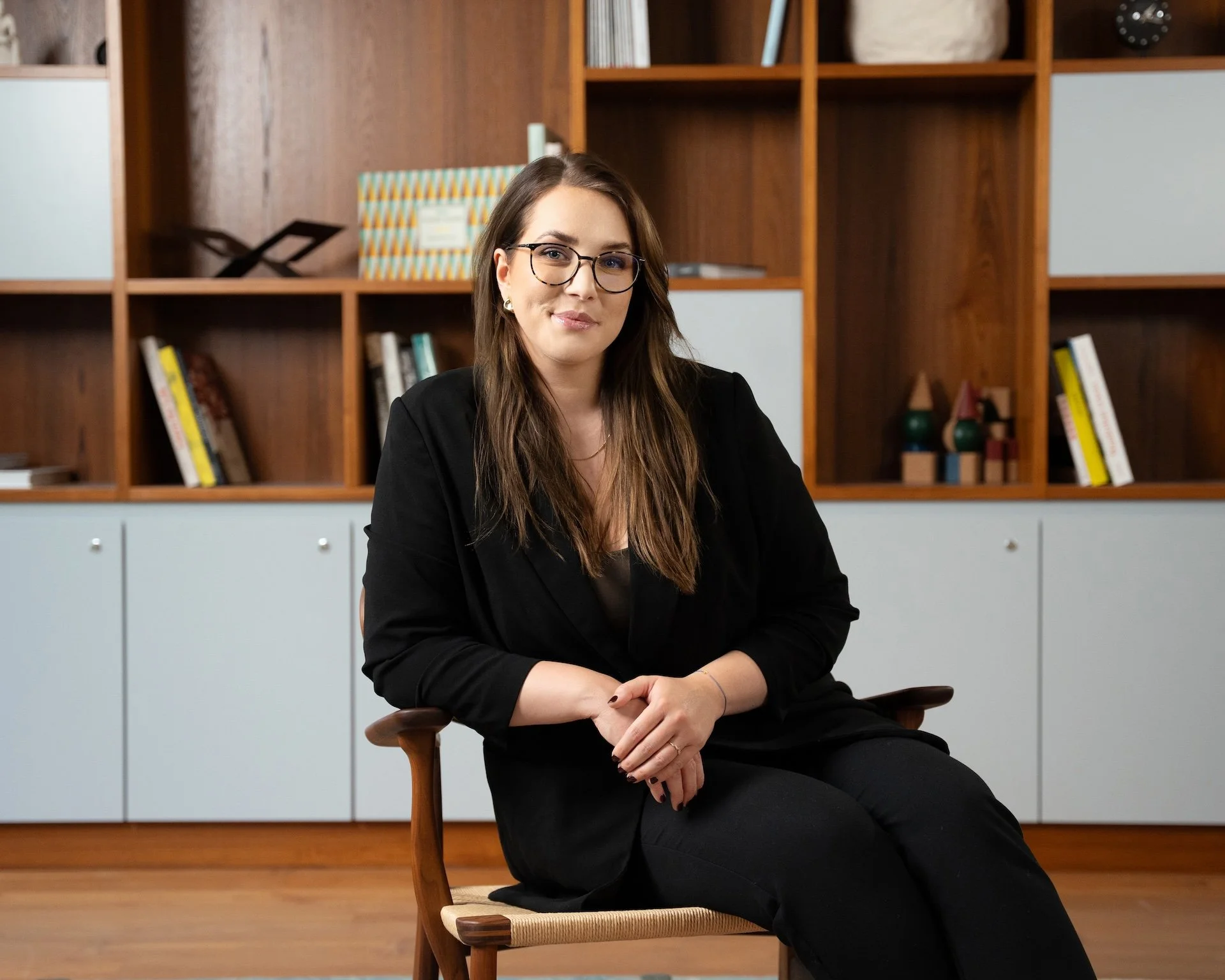 A woman with brown hair, glasses, and earrings, wearing a black blazer, sitting on a wooden chair in a room with wooden shelves and cabinets behind her, some of which contain books and decorative items.