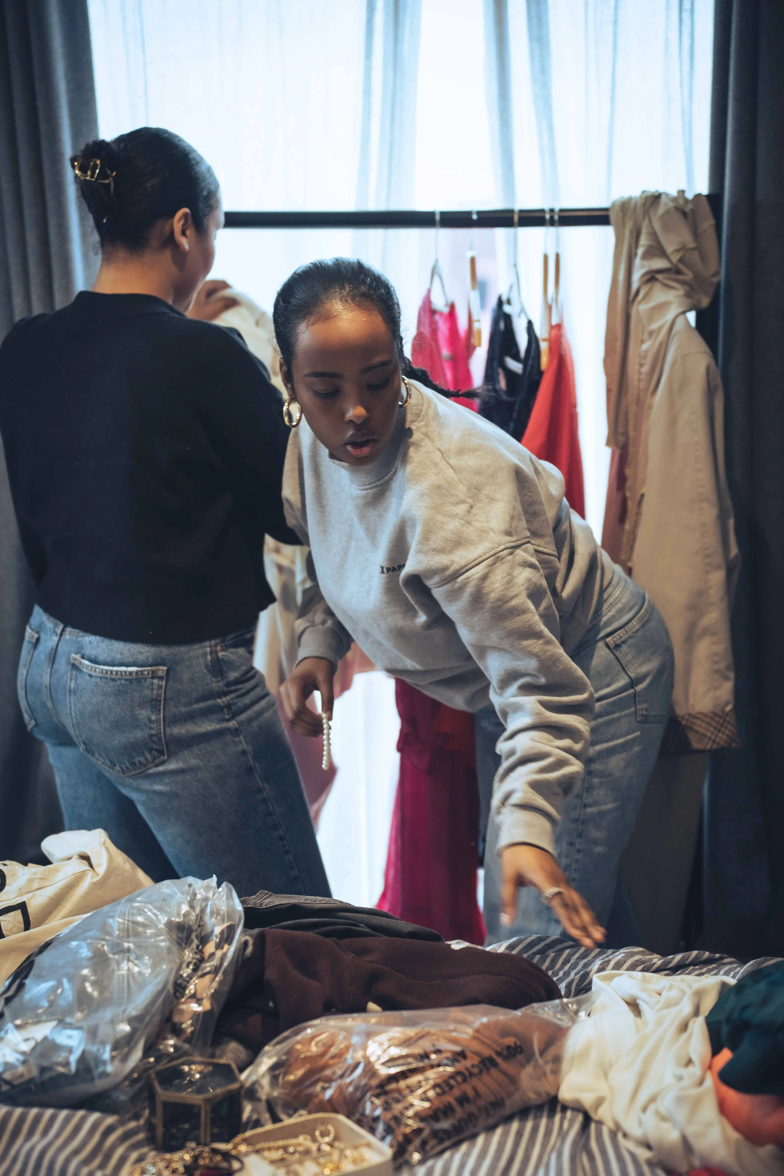 Two women organizing clothing on a bed with various jewelry and accessories scattered around, in a room with a window and clothing hanging in the background.