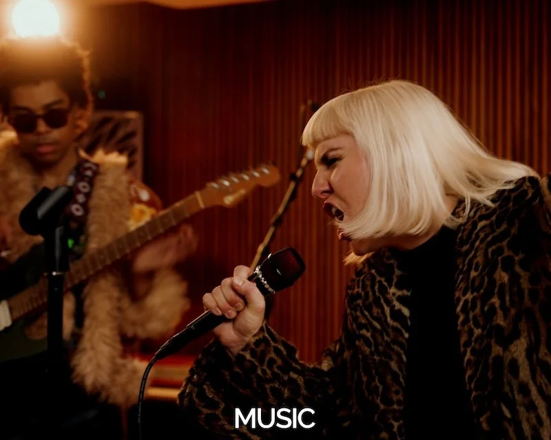 A woman with platinum blonde hair singing into a microphone while a person with sunglasses and a fur coat plays an electric guitar in the background.