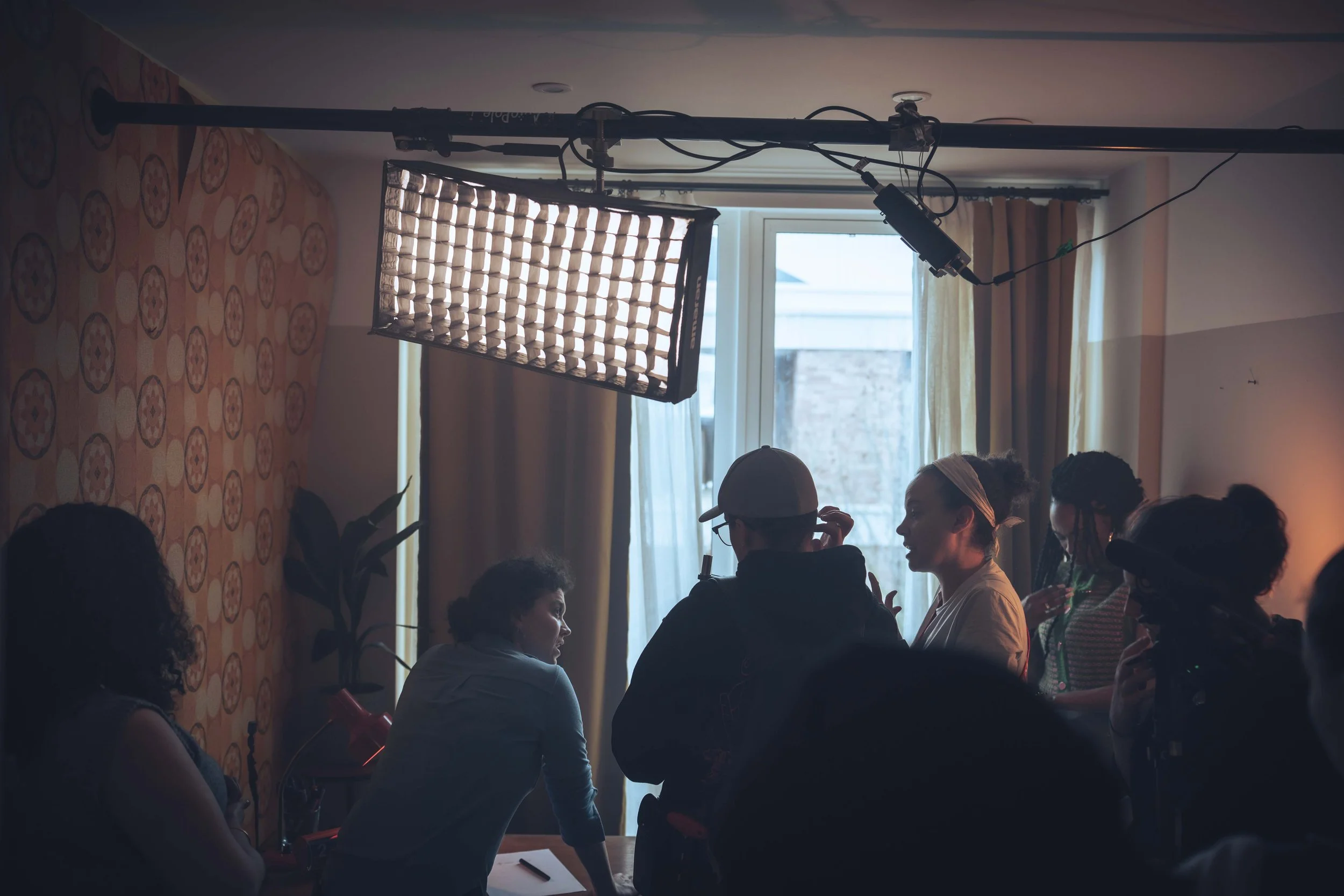People seated around a conference table engaged in discussion, with a bright window behind them and studio lighting equipment overhead.