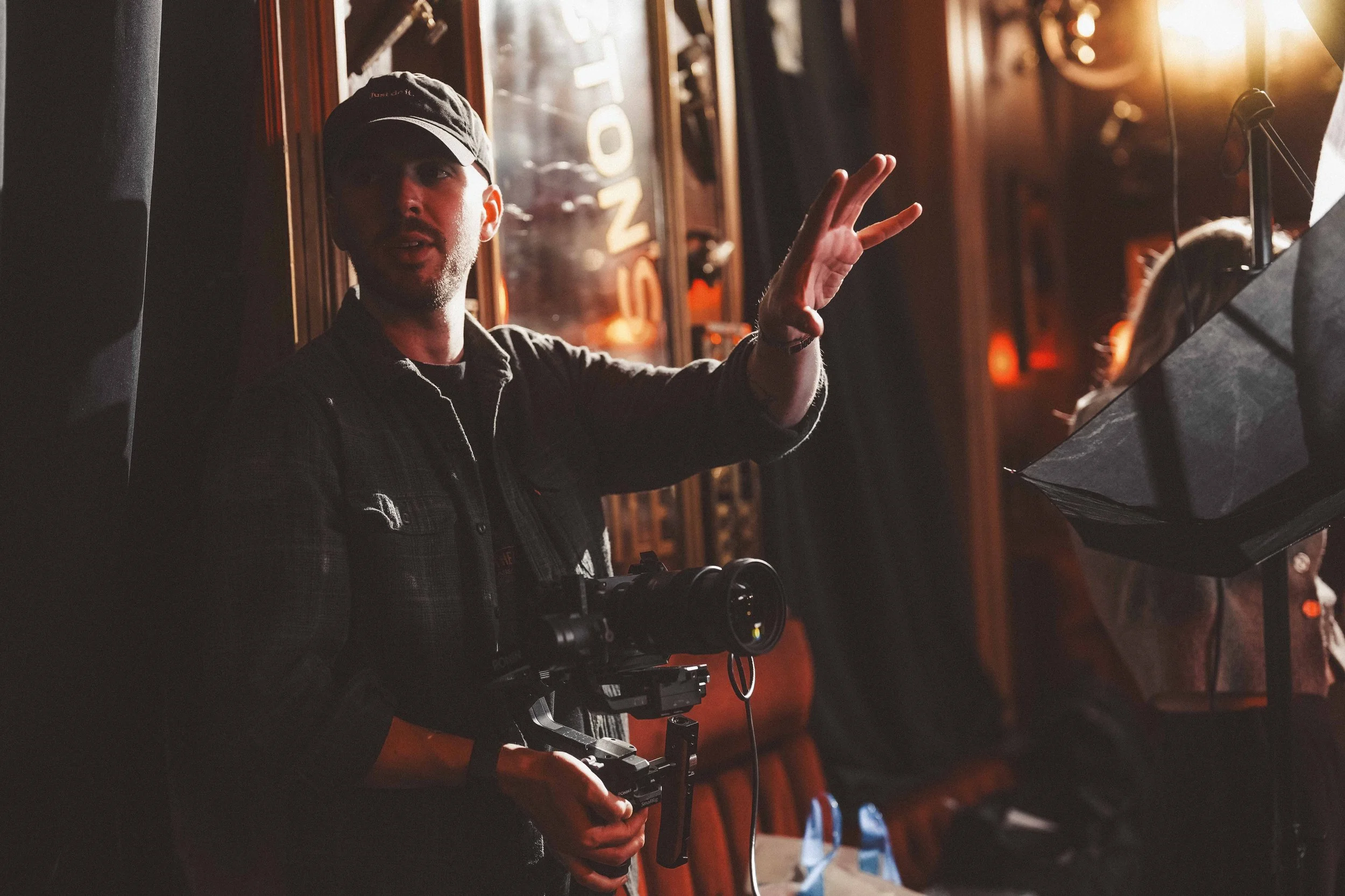 A man wearing a black cap and dark clothing is speaking or giving instructions, holding a camera stabilizer, in a dimly lit indoor setting with wooden details and warm lighting.