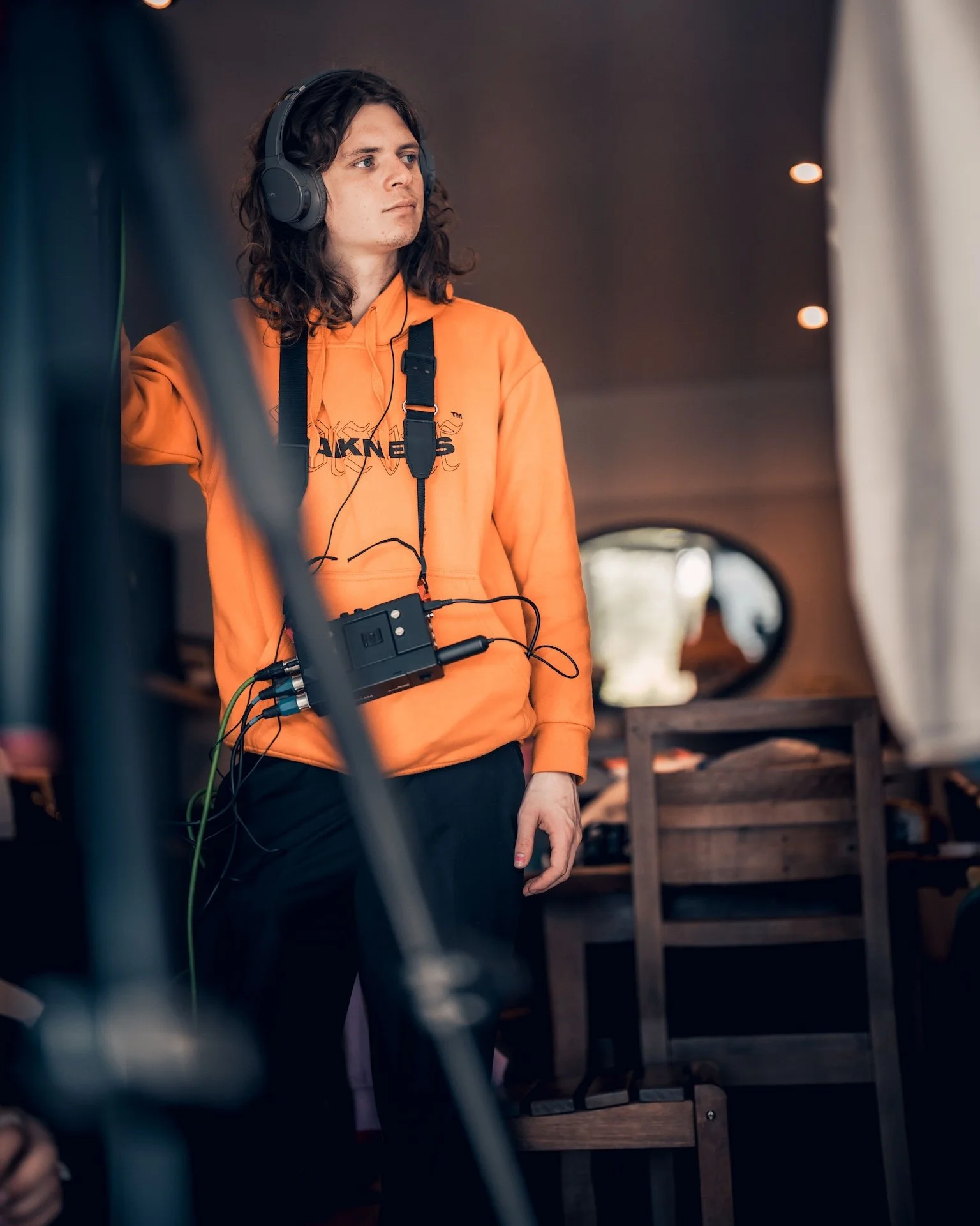 Young man with long curly hair wearing a bright orange hoodie, black headphones, and a communications device around his neck, standing indoors near wood furniture.