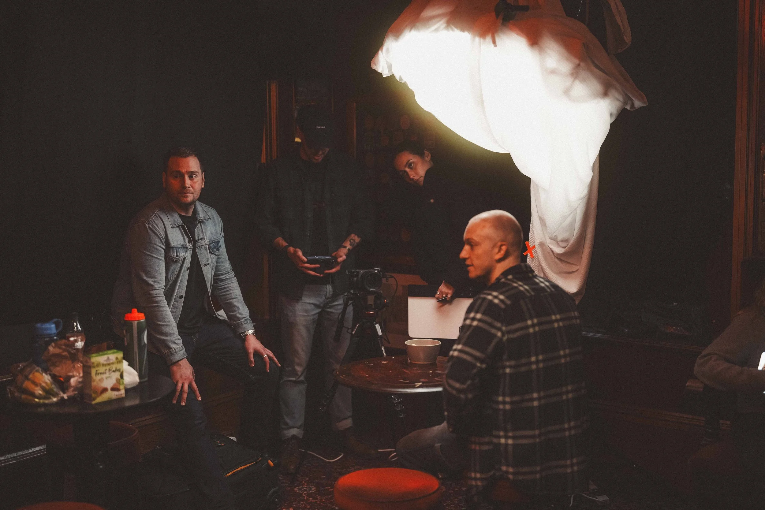 A group of people are gathered in a dimly lit room, with some standing and others sitting around a table. There are men and women, and the scene appears to be a casual social or work gathering. One person is holding a camera, and snacks and drinks ar