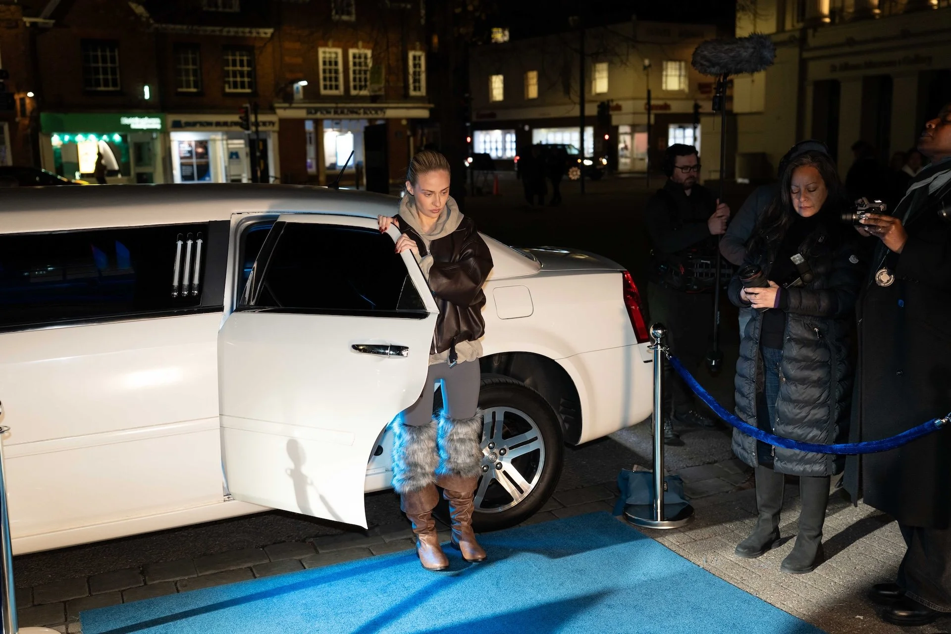 Woman with blonde hair, wearing a black leather jacket, gray hoodie, and furry brown boots, standing next to a white limousine at night, with a film crew recording her.