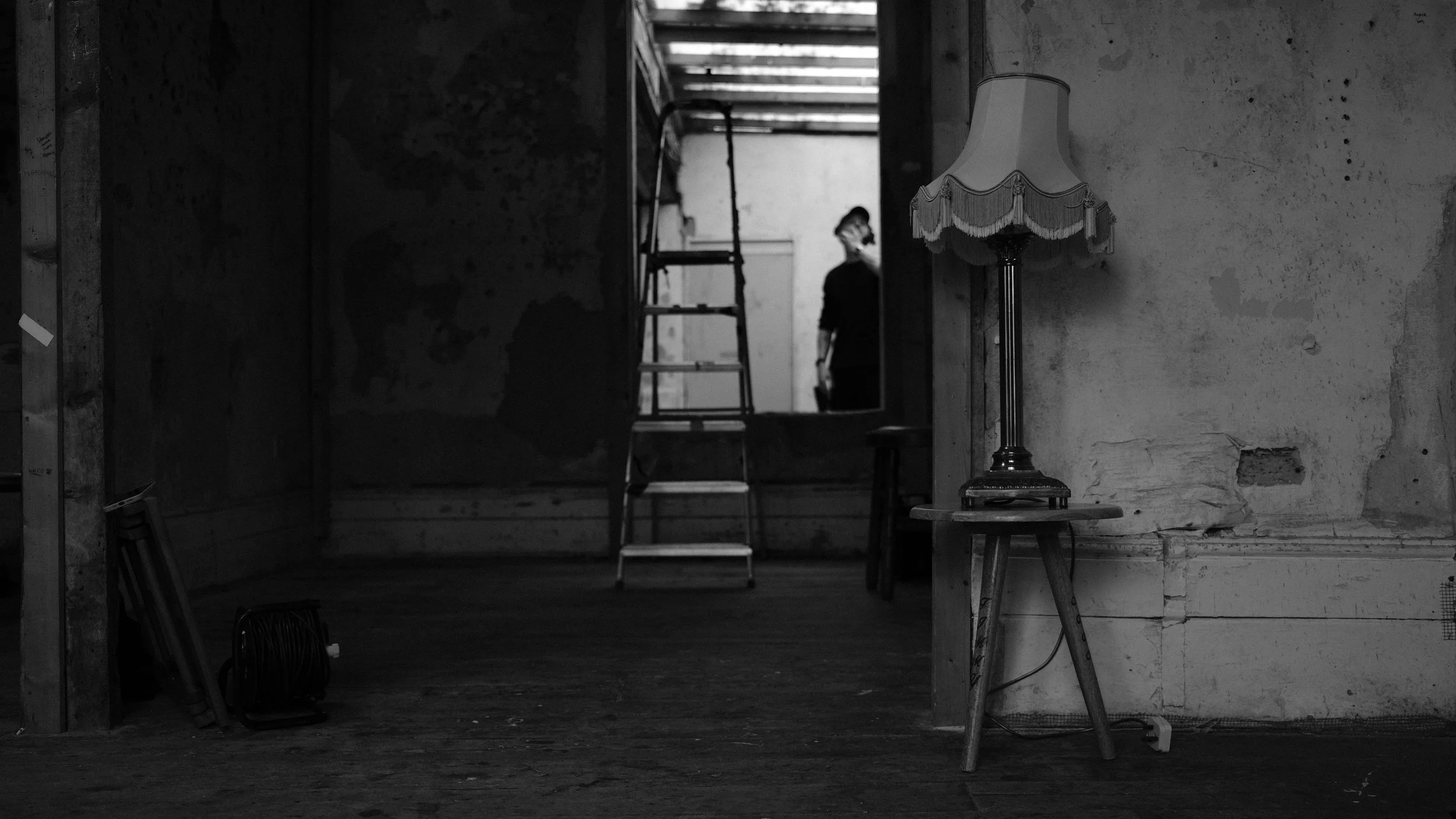 An empty room with a vintage lamp on a small stool, a ladder leading to an opening above, and a person standing in the background near a wall with peeling paint, in black and white.
