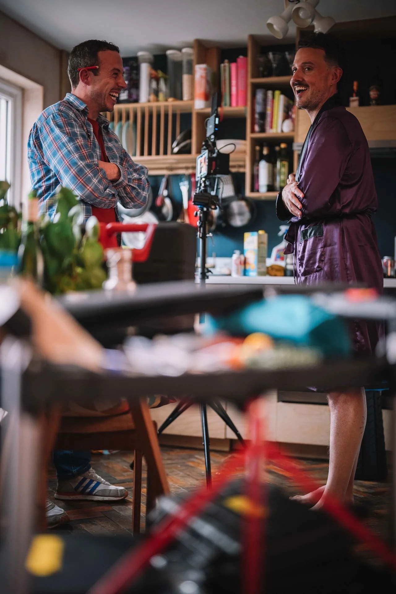 Two men in a kitchen, laughing and smiling while talking. One man is wearing a plaid shirt, the other is in a dark robe. The photo is taken from a low angle with some kitchen items in the foreground.