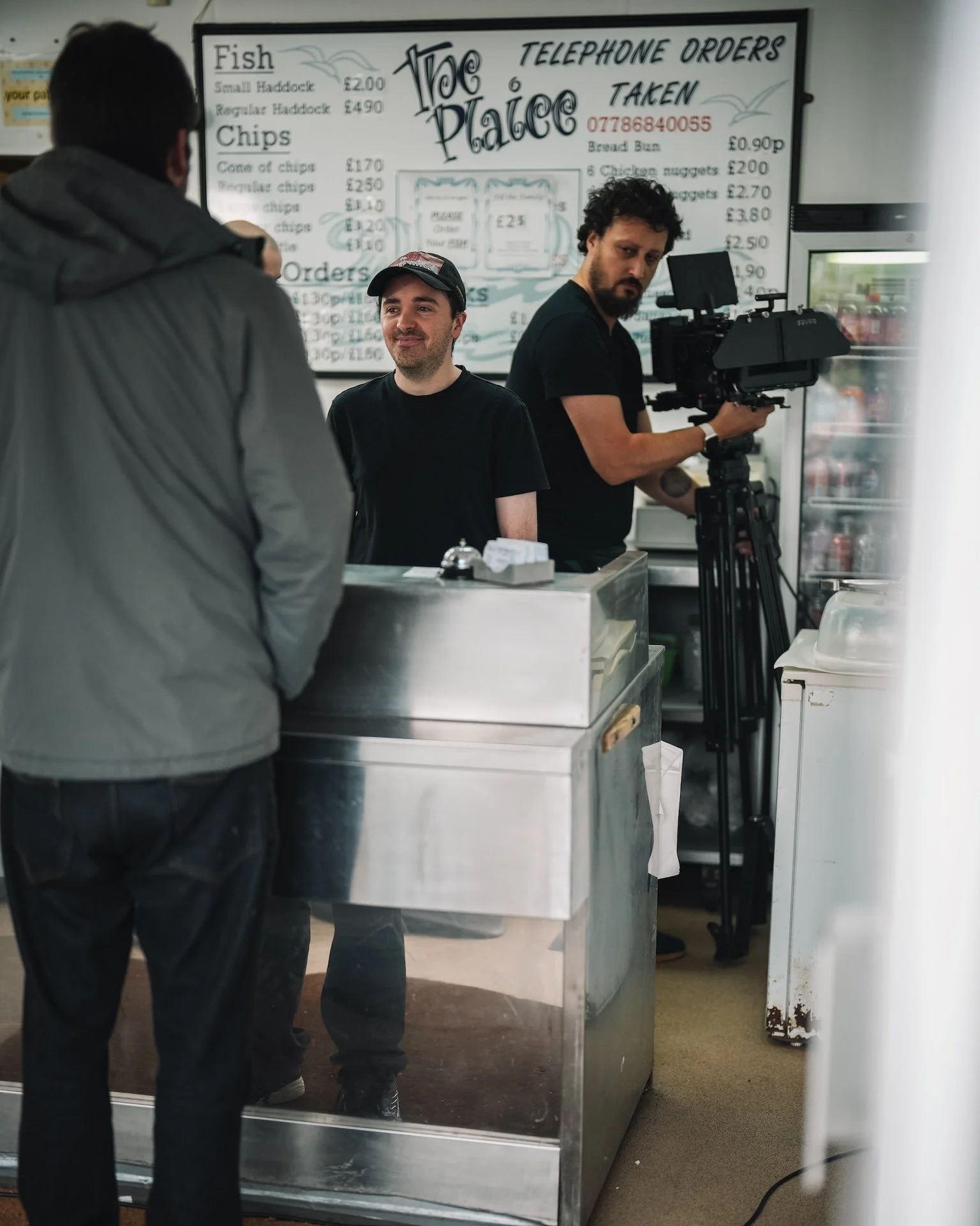 A scene inside a fish and chips shop with two men working behind the counter, one with curly hair and a beard and the other with light facial hair, and a customer wearing glasses and a gray jacket. A film crew with camera equipment is filming the sce