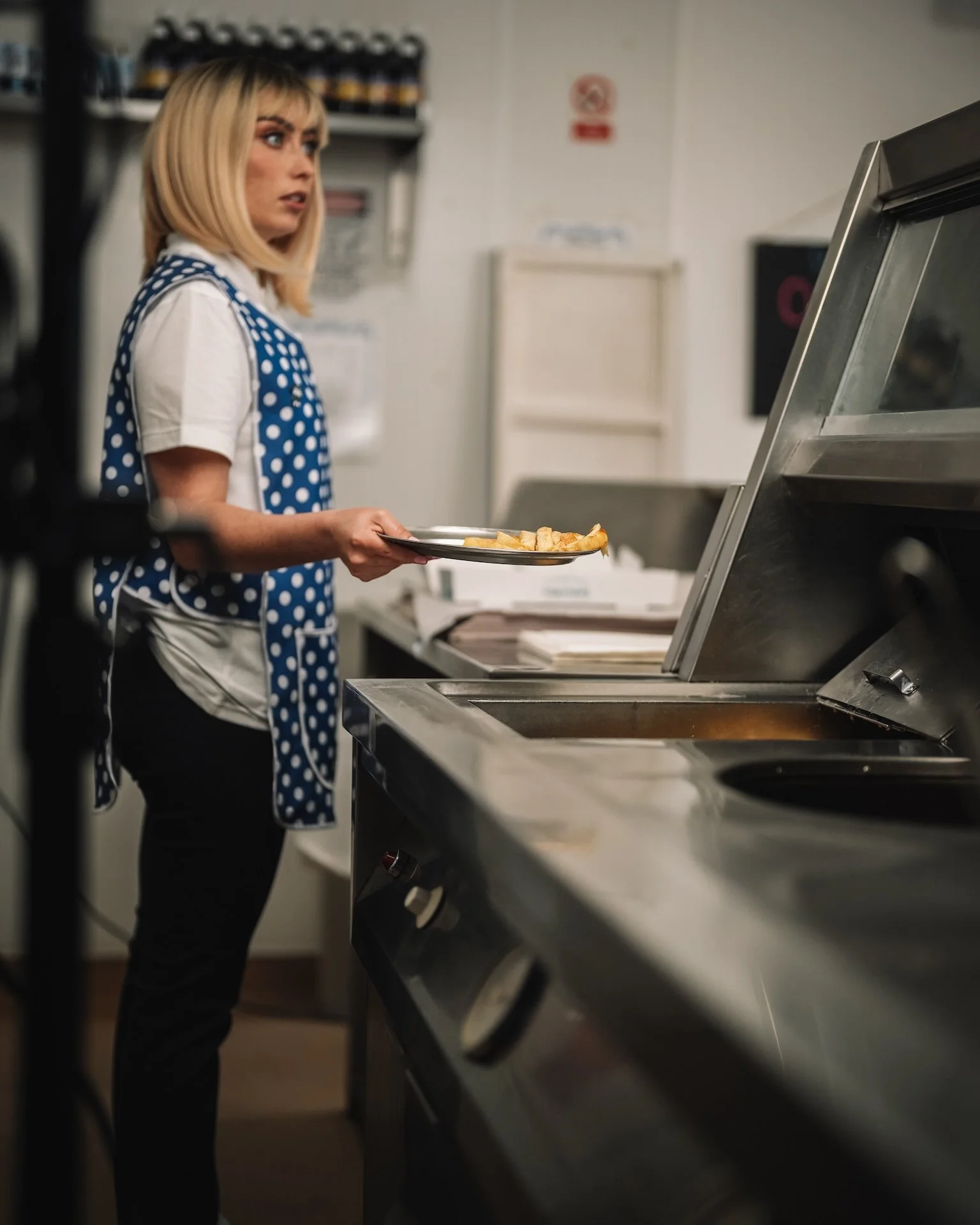 A woman with blonde hair, wearing a white shirt and a blue polka dot apron, is holding a plate of food near a commercial kitchen stove.
