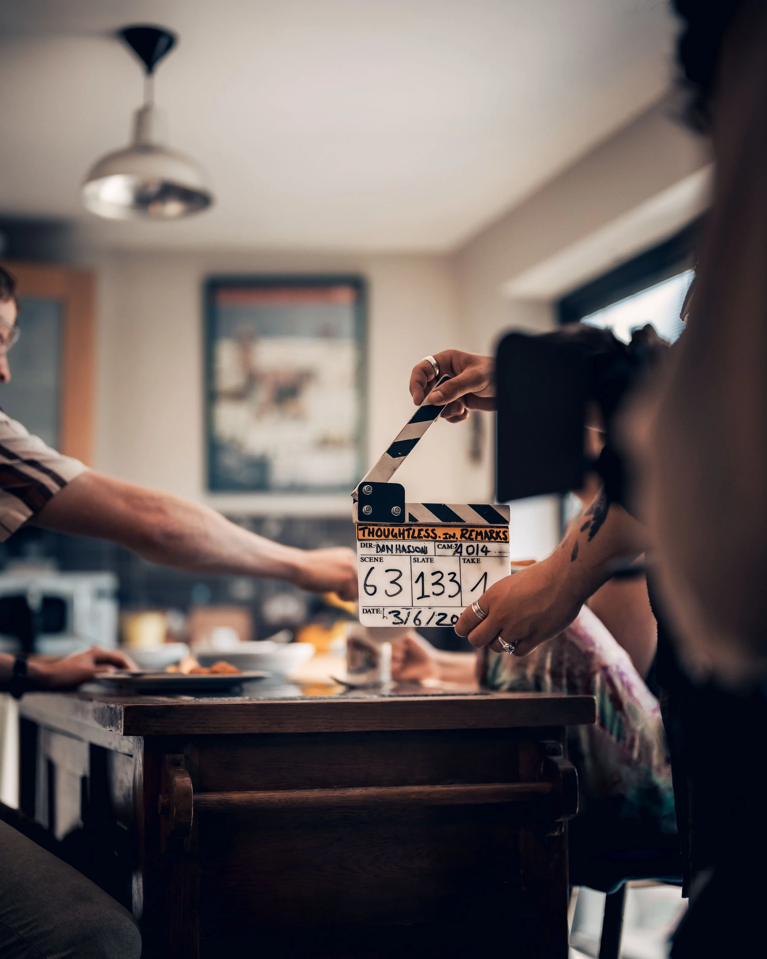 People sitting around a wooden table, one person holding a clapperboard labeled 'Thoughtless in Remarks,' with scene and take numbers, in a room with framed artwork and a window.