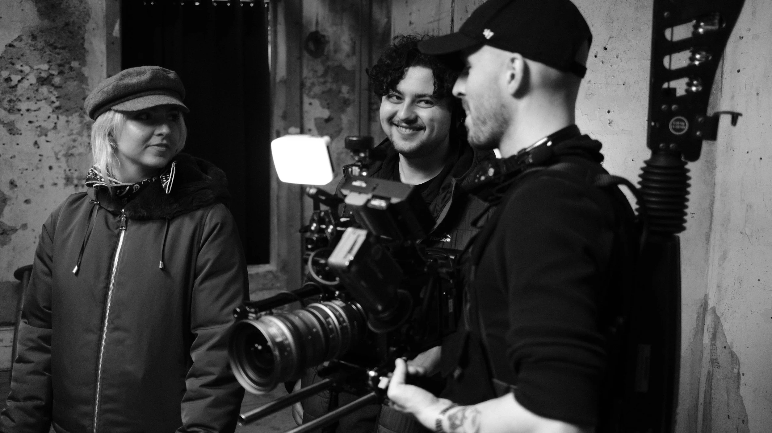 Three people on a film set: a woman wearing a beret, a smiling man with curly hair, and a man with a cap operating a professional camera.