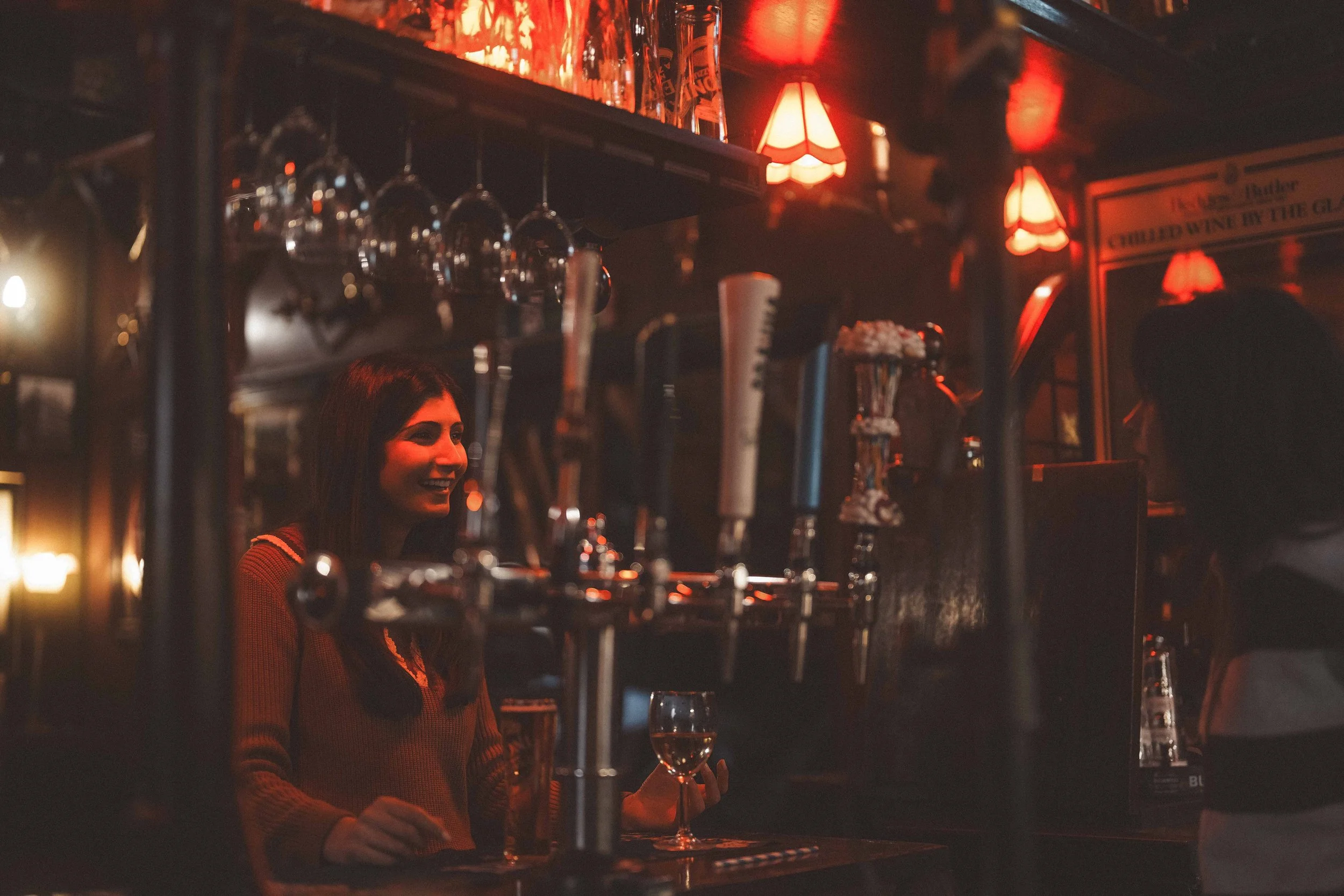 A woman smiling and holding a glass of wine behind a bar with red lighting, shelves with liquor bottles, and beer taps.