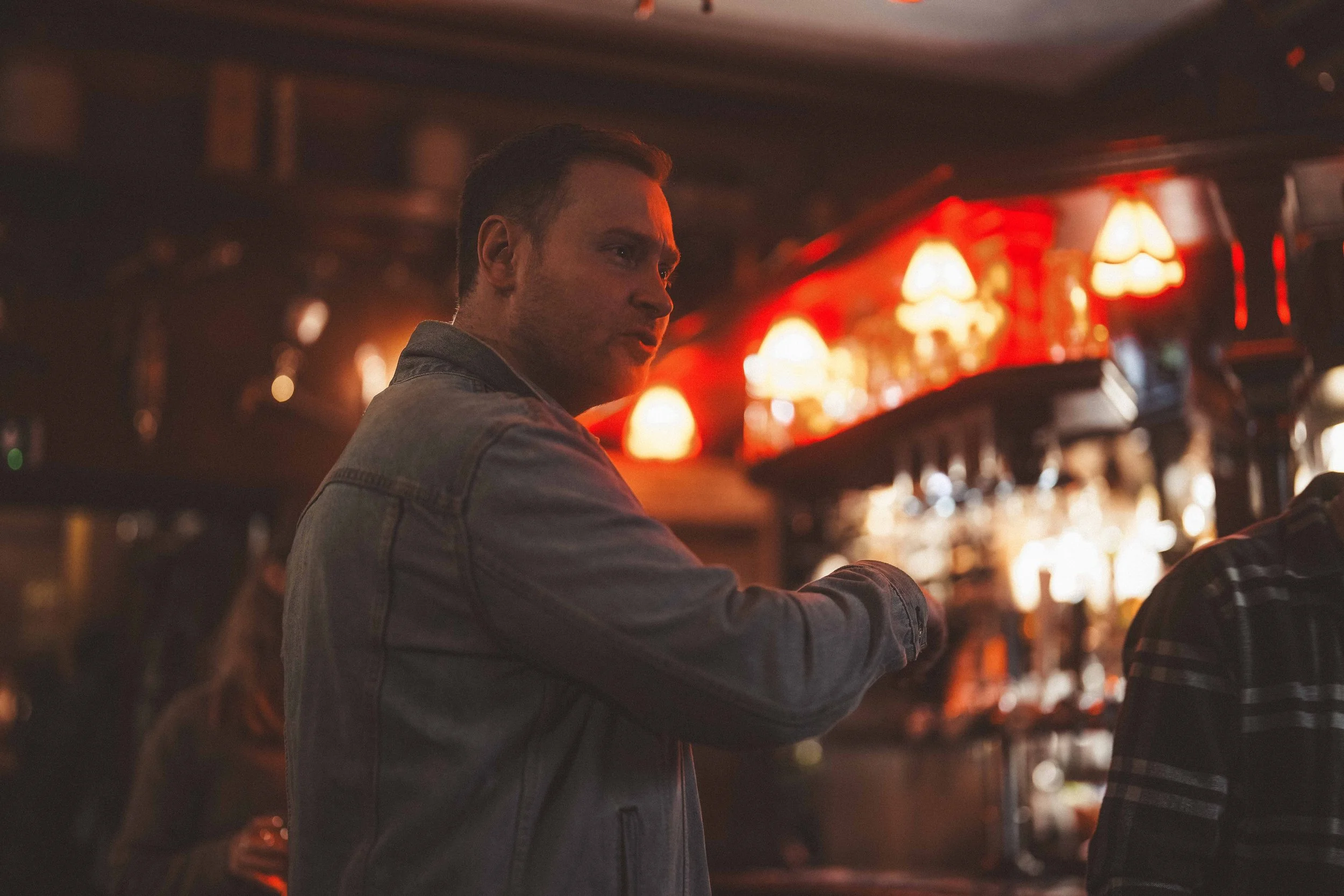 A young man with short dark hair is sitting at a bar with warm red and orange lighting. He is wearing a denim jacket and appears to be engaged in a conversation or deep in thought. The bar is dimly lit with decorative lamps and bottles in the backgro