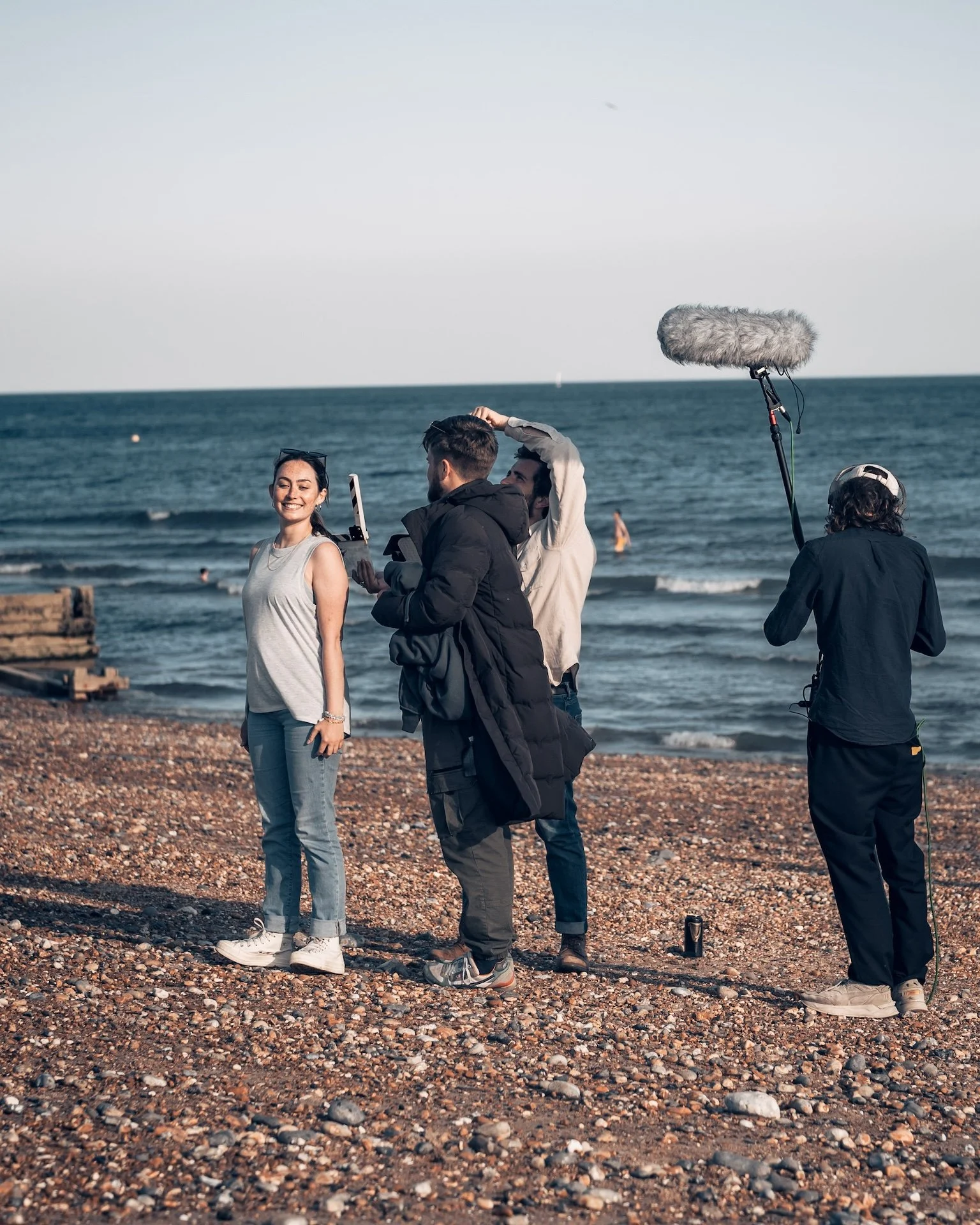 A woman smiling on a pebble beach with a film crew recording her, ocean waves in the background, crew members filming and holding audio equipment.
