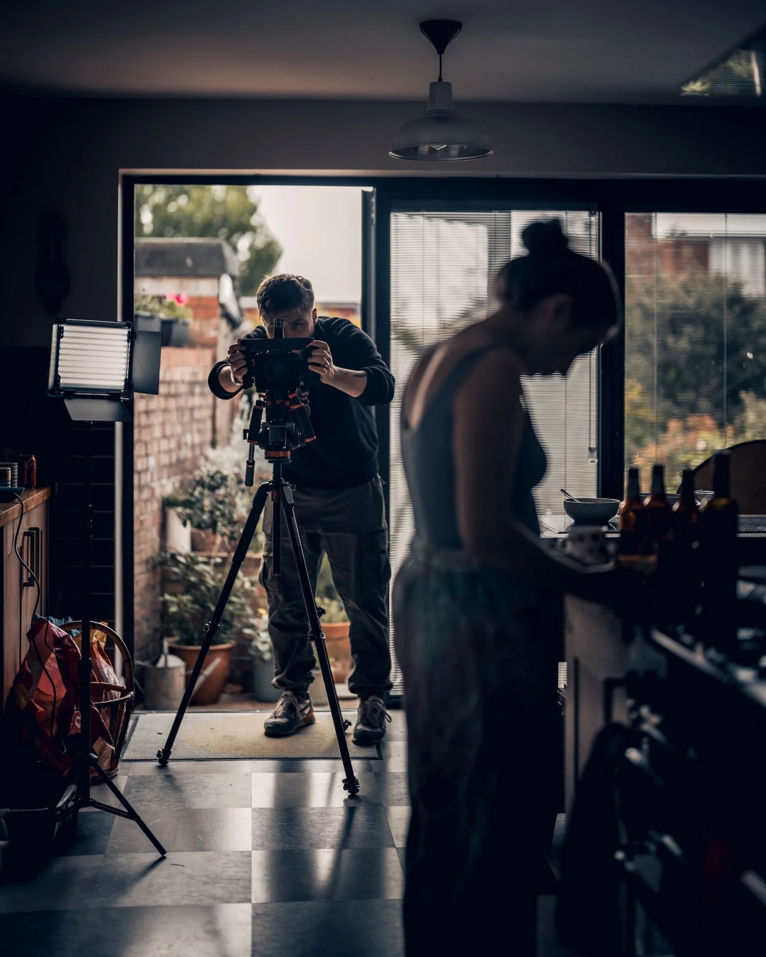 Person setting up a camera on a tripod in a doorway, with another person in the foreground, and outdoor plants visible through the open door.