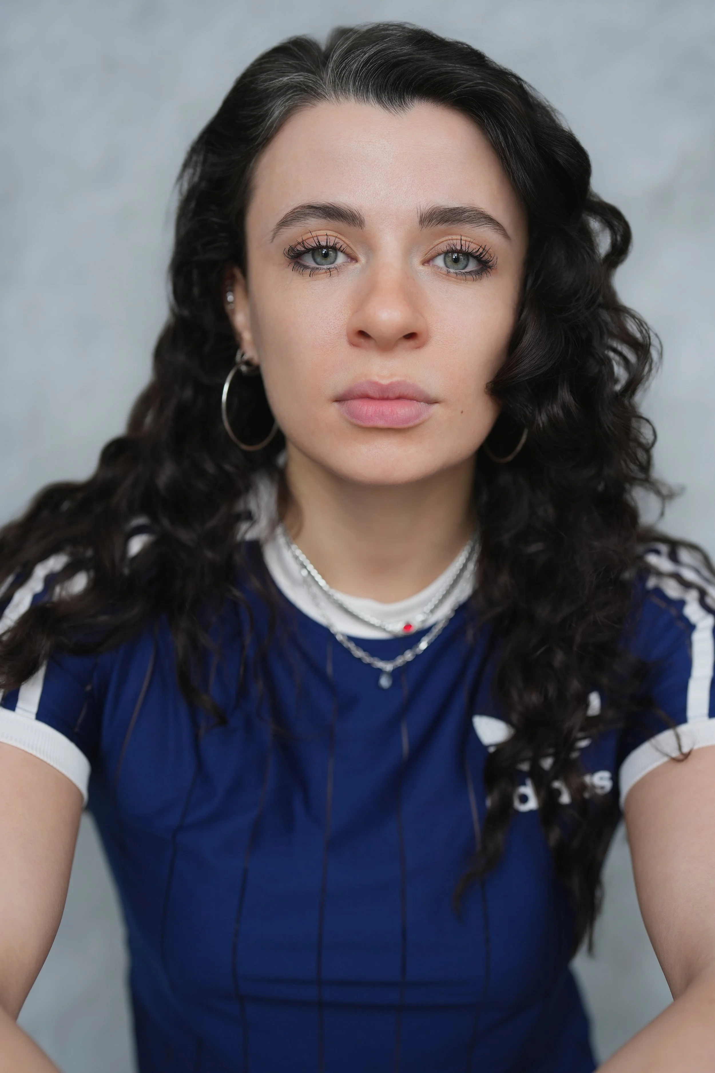 Close-up of a young woman with long, curly black hair, wearing large hoop earrings, layered necklaces, and a blue sports jersey with white accents, against a blurred gray background.