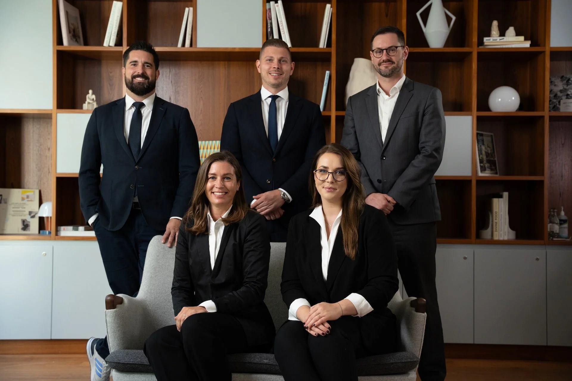 Group photo of five business professionals, three men and two women, dressed in formal suits, posing indoors in front of a wooden bookshelf with decorative items and books.