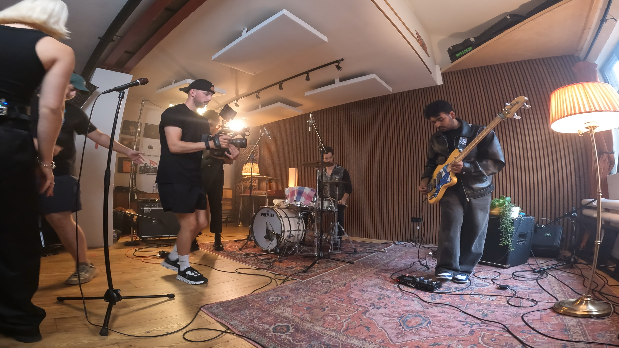 A band of five musicians rehearsing indoors, with a camera person filming them, a drummer behind a drum set, a guitarist wearing a leather jacket playing electric guitar, and others around, with lighting and sound equipment present.