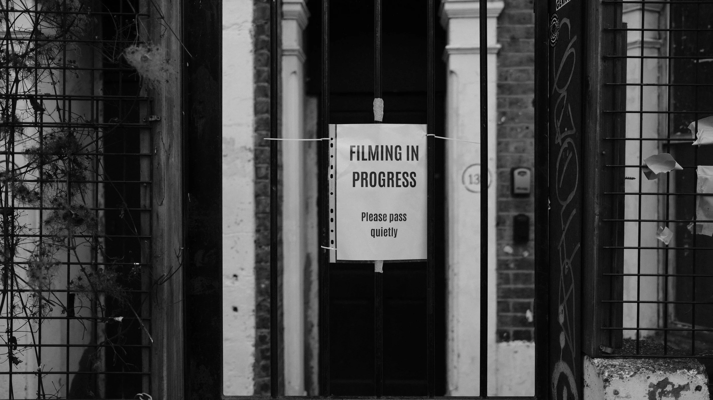 A black and white photo of a metal gate with a sign that reads 'Filming in Progress, Please pass quietly.' The gate is flanked by brick and wood structures and some overgrown plants.