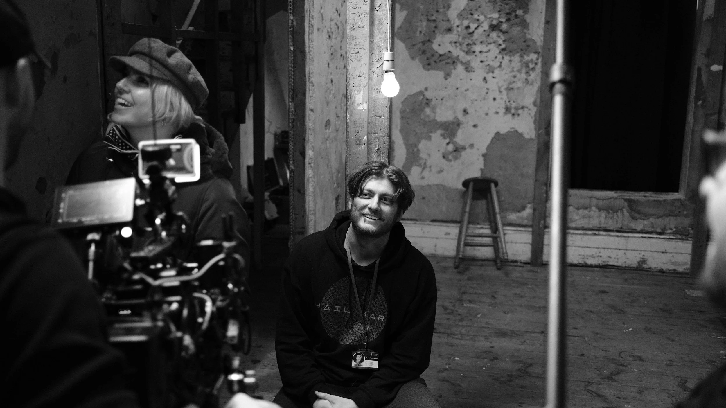 A woman with a hat and a smiling man with a badge sitting on the floor in a rustic, unfinished room with peeling walls and a stool.