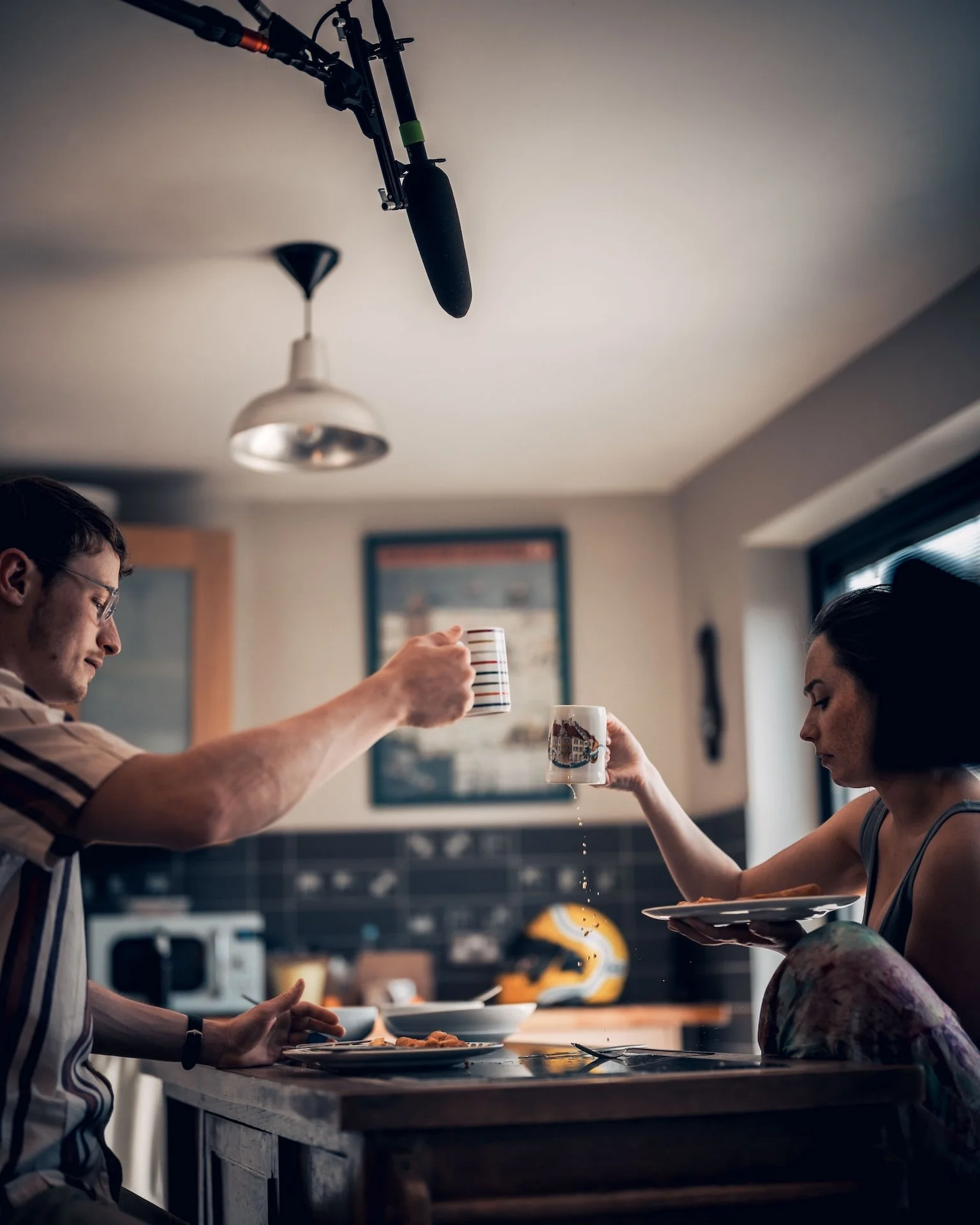 Two people sharing coffee at a kitchen counter, with one person passing a mug to the other. A man is on the left side, and a woman is on the right. The scene has a casual and warm atmosphere.