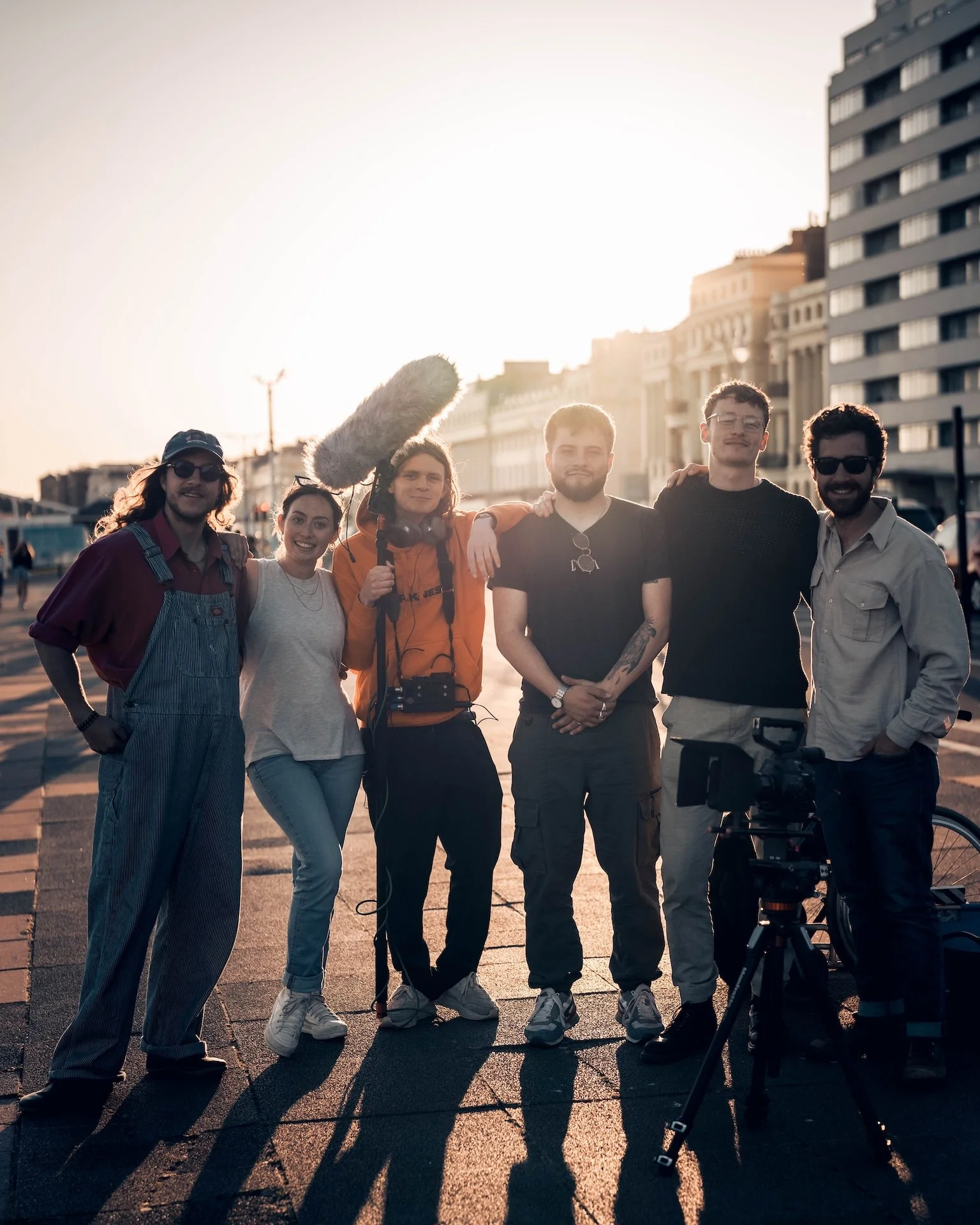 Group of six people standing on a city street at sunset with film equipment, smiling and posing for a photo.