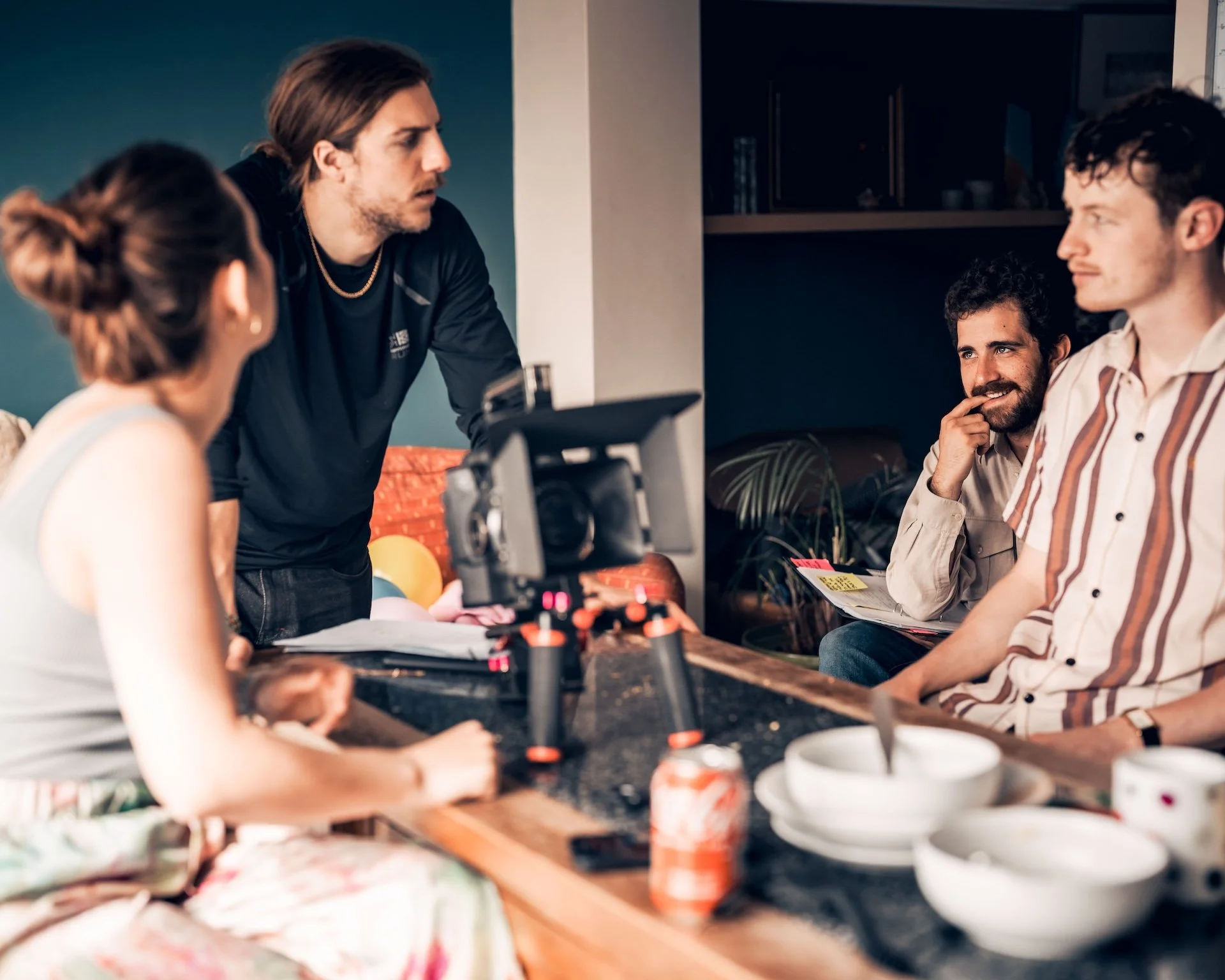 Four people sitting at a kitchen table during a discussion, with a woman operating a camera and balloons in the background.