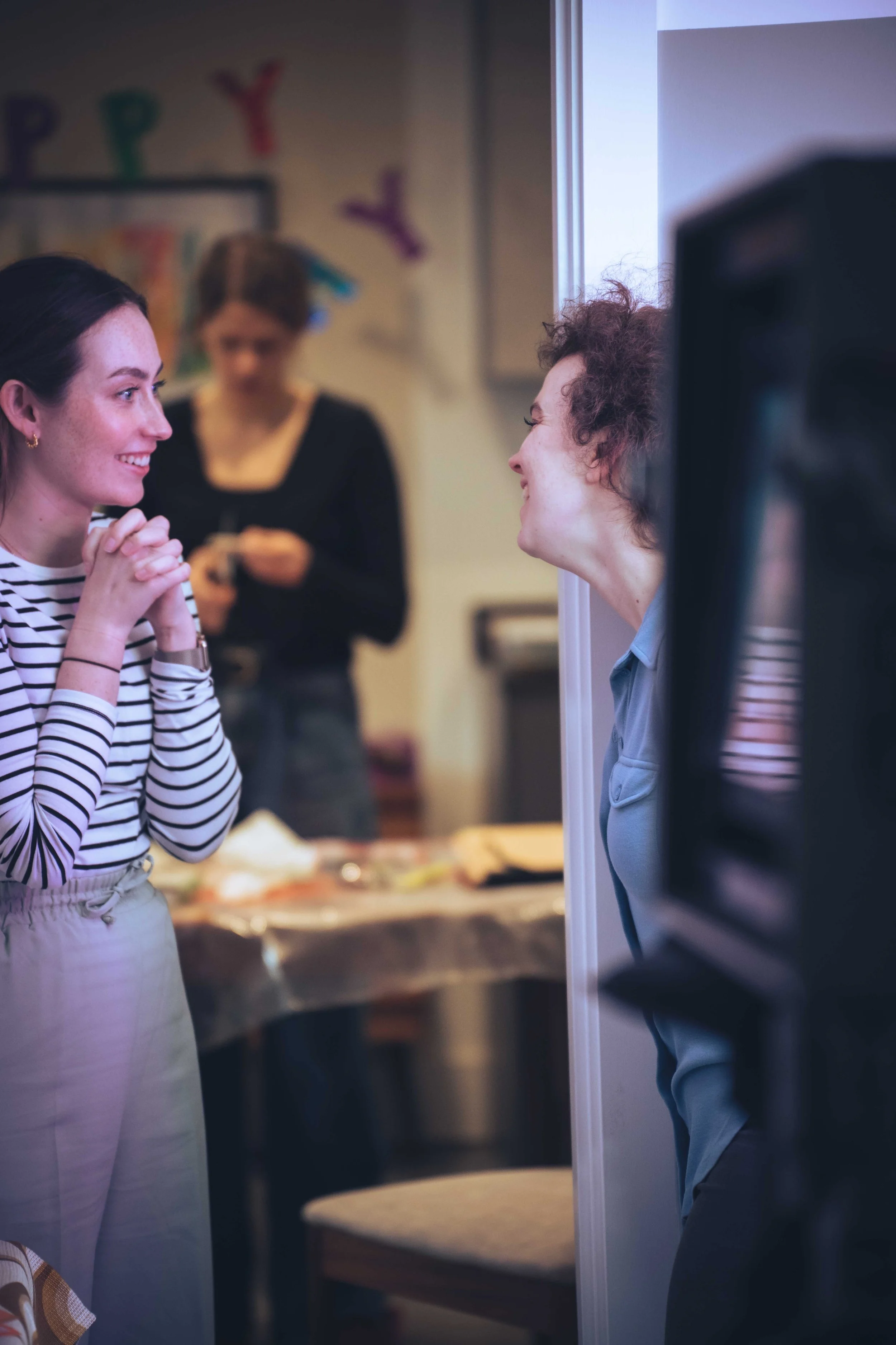 A woman and a man are facing each other, smiling and engaging in conversation in a room with a window. The woman is wearing a striped shirt and has her hands clasped together, while the man is leaning on a wall.