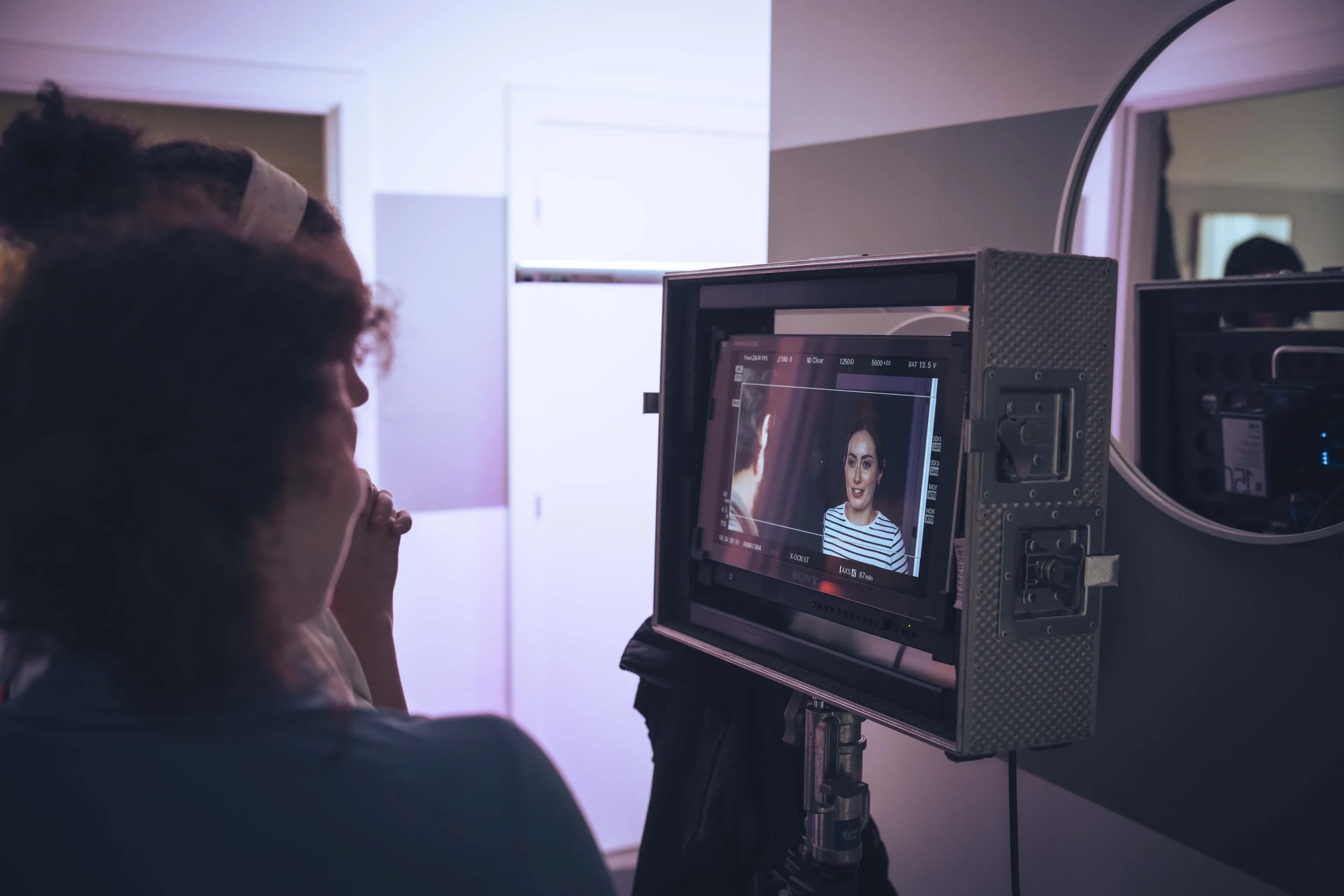 A person on set during a photoshoot or video shoot looks at a monitor displaying a woman in a striped shirt being filmed.