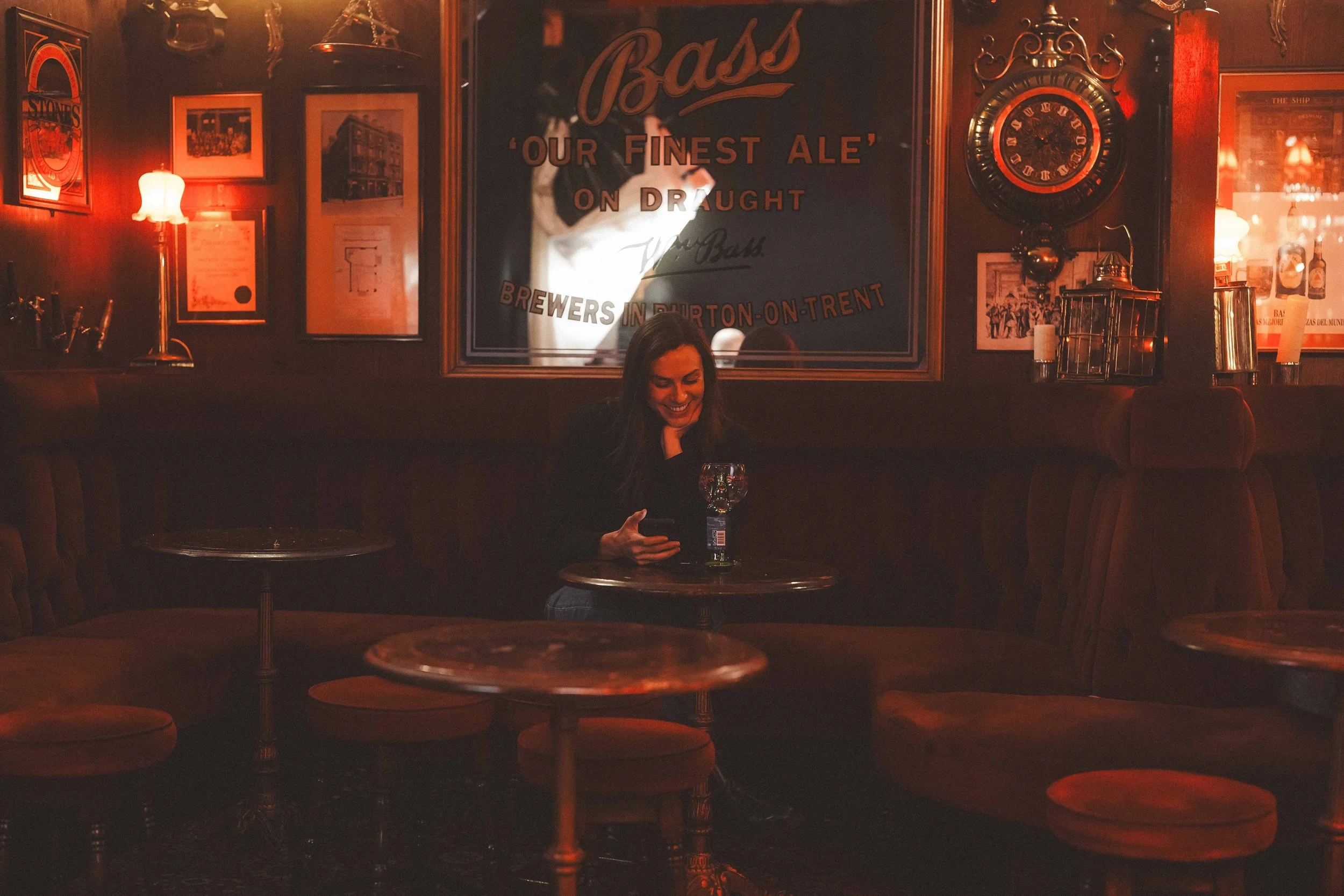 A woman sitting alone at a small round table in a dimly lit pub or bar, looking at her phone and smiling. The background features vintage decor including framed pictures, a large wall clock, warm lighting, and a sign that reads 'Bass Our Finest Ale o