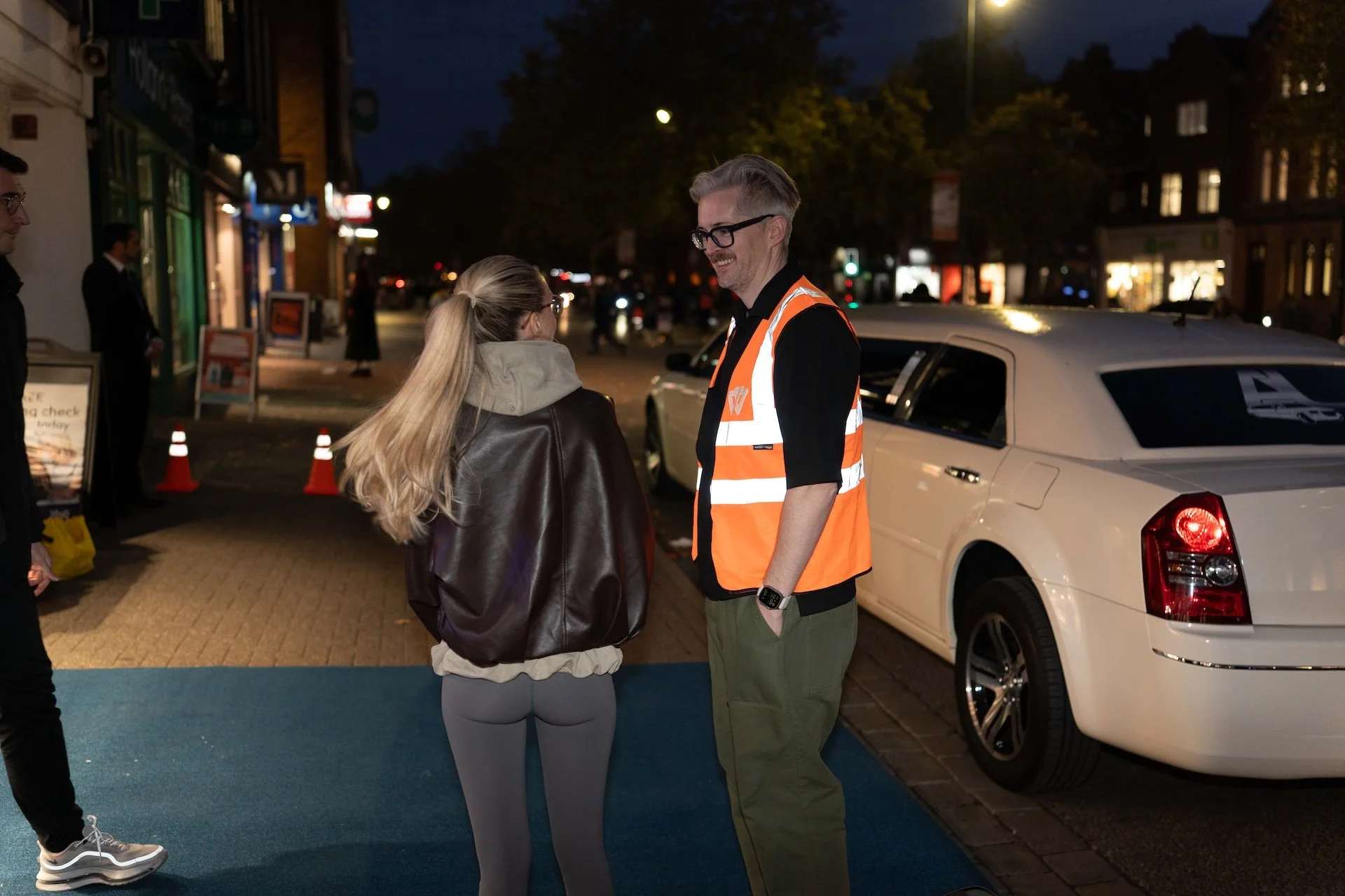 A man wearing glasses and an orange safety vest talking to a woman with long blonde hair pulled back, wearing a leather jacket, on a city street at night. There is a white car parked nearby, and several pedestrians and shops are visible in the backgr