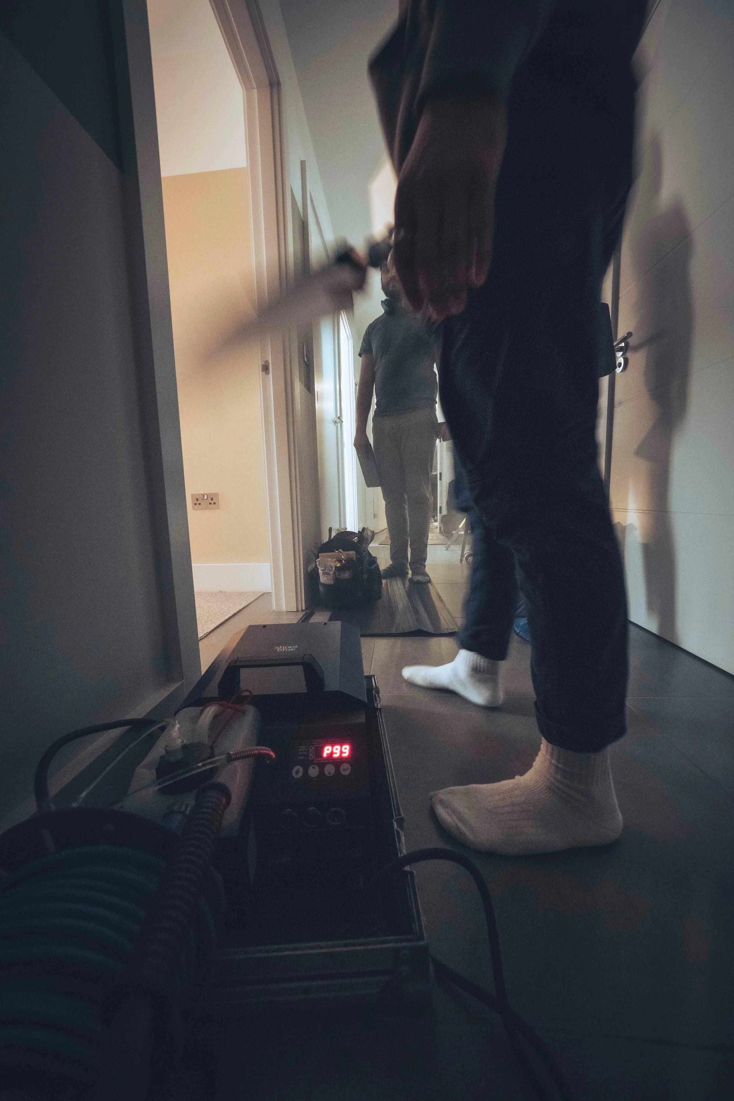 Indoor scene with a person standing in a hallway, holding a paper, and a dog in the background near a door. In the foreground, there is a device on the floor displaying 'P99'.