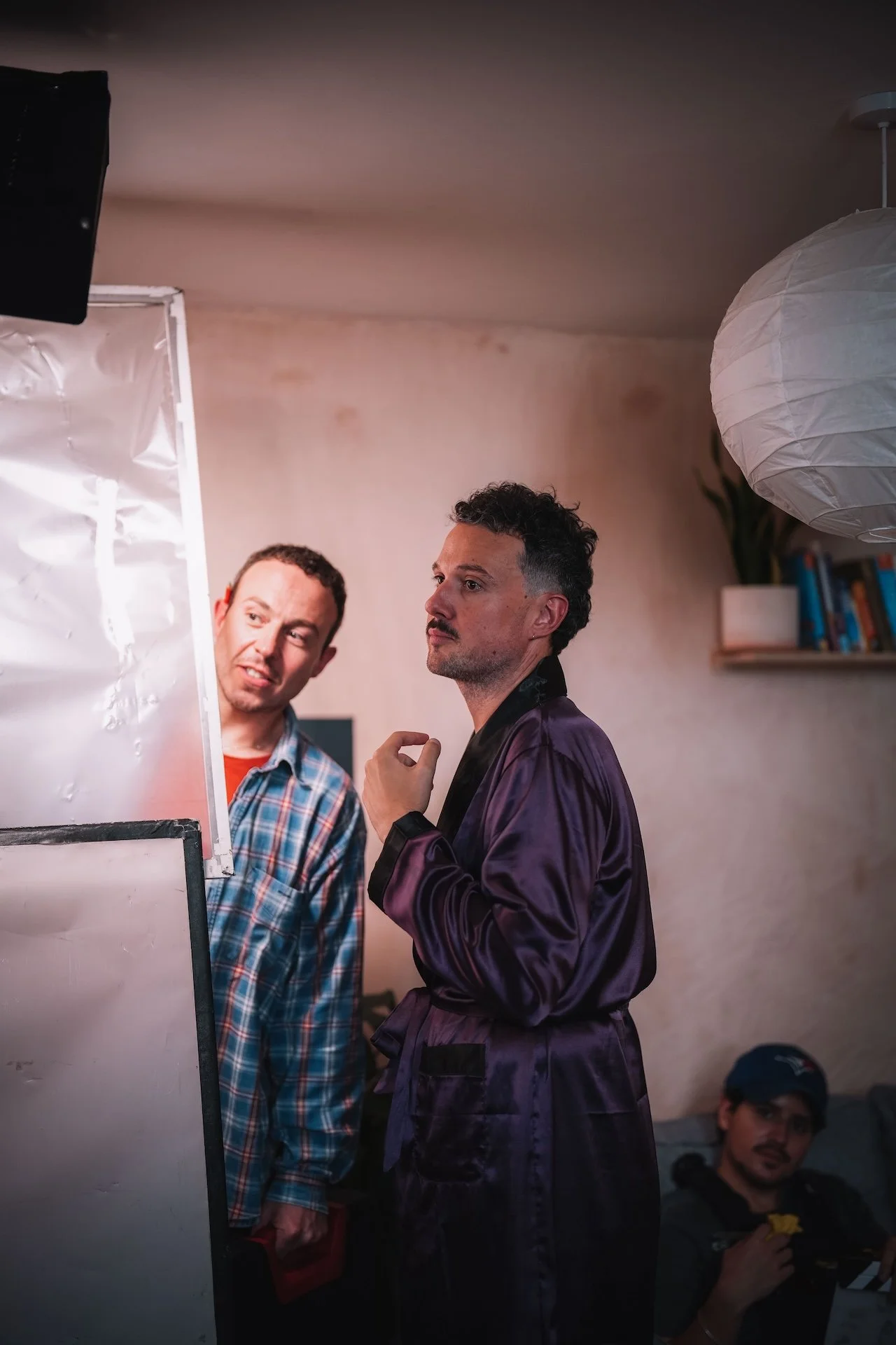 Two men engaged in conversation indoors, one wearing a plaid shirt and the other in a shiny purple silk robe; a third man sitting on a couch in the background.