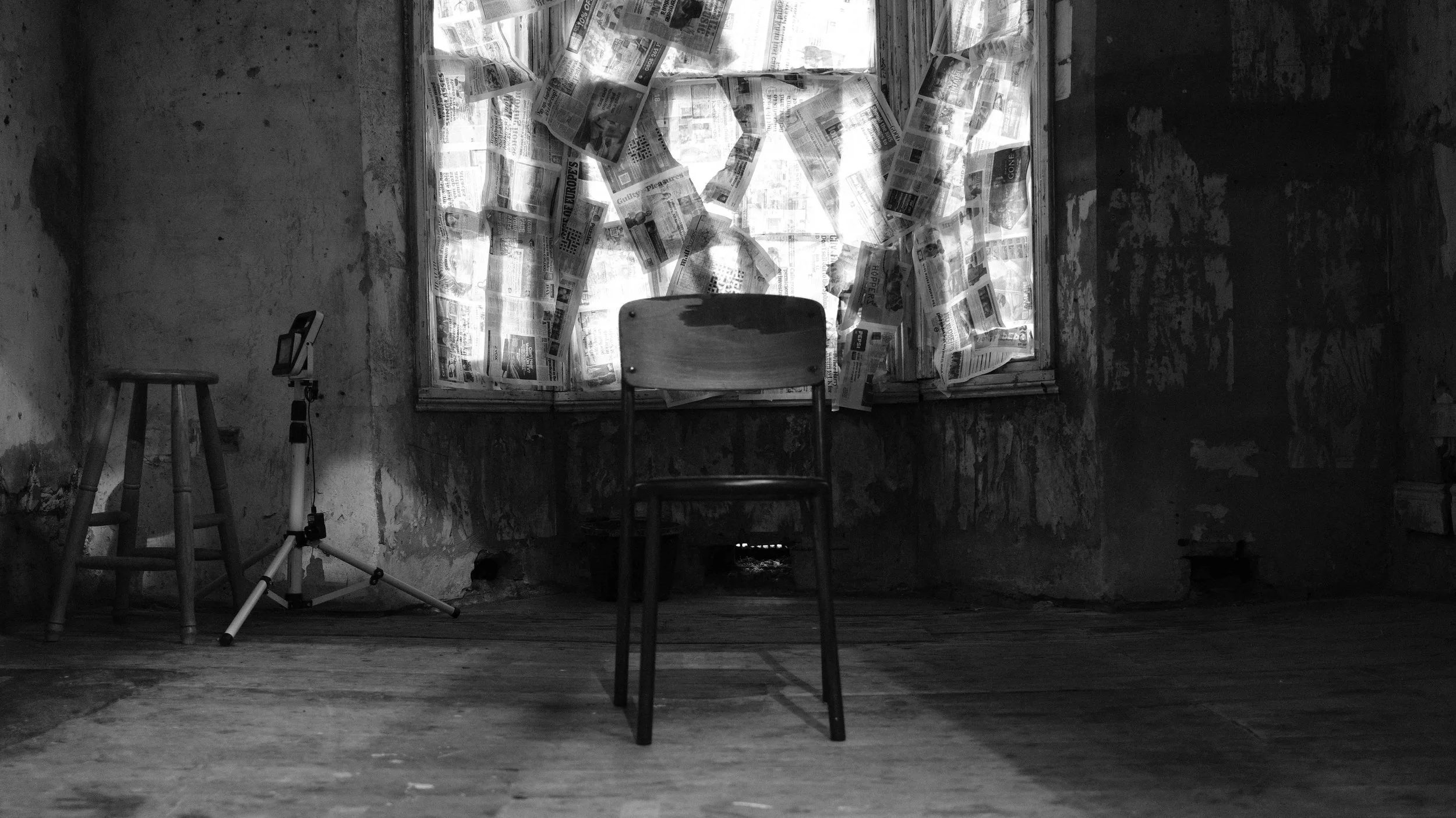 A black and white photo of an empty room with a wooden floor and a window covered in crumpled newspapers. In the center of the room, there is a wooden chair, and to the left, an stool and a tripod with a mounted device.
