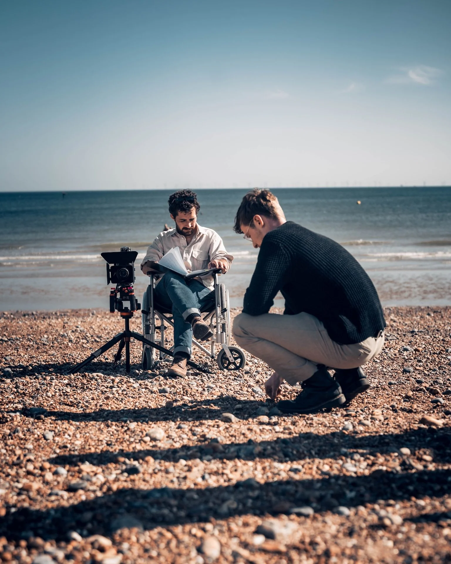 Two men on a pebbled beach with the ocean in the background. One man is sitting in a wheelchair with a camera and papers nearby, while the other man is crouched down, possibly adjusting something on the ground.