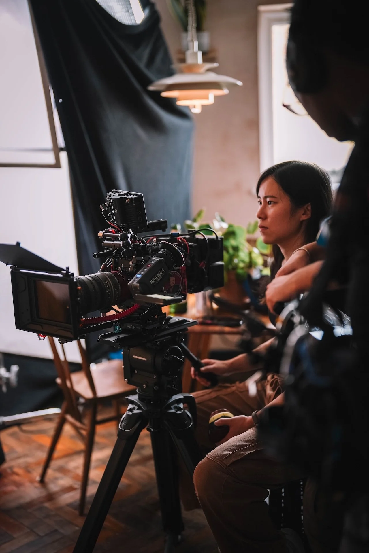 A woman is sitting in front of a camera with a crew member standing nearby. The scene is set indoors with warm lighting, plants, and a window in the background.