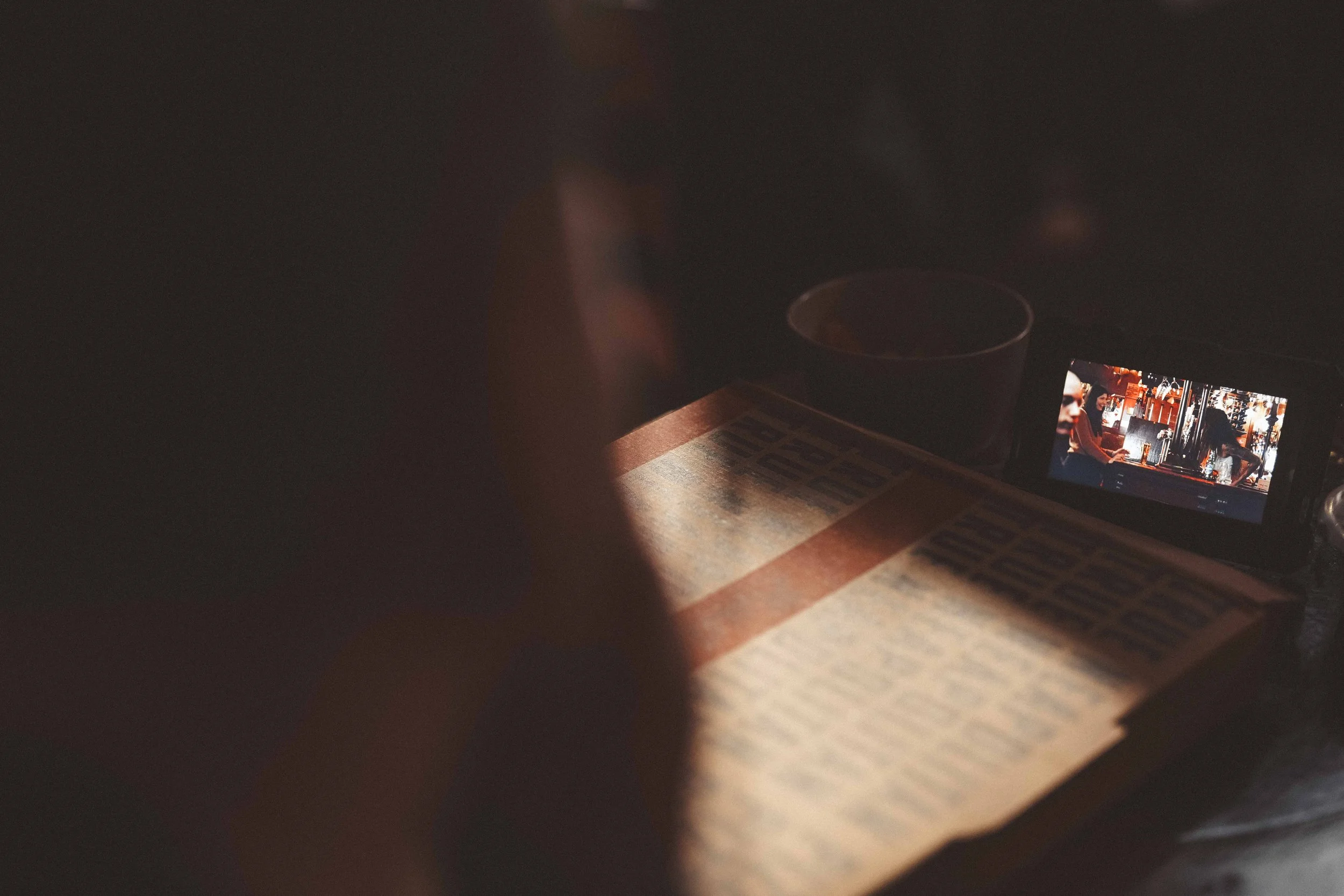 Photograph of a cellphone displaying a photo of two people at a bar, with a drink and menu on a table, taken through a narrow opening in darkness.