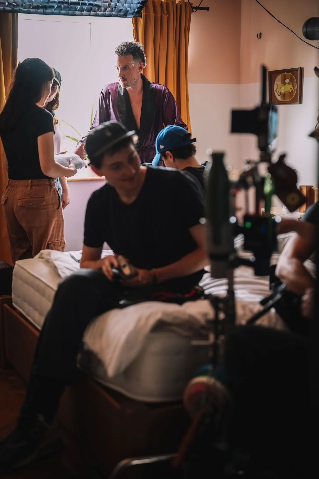 Filming crew in a bedroom with a man in a satin robe, two women, and a person in a hat sitting on the bed, with filming equipment around.