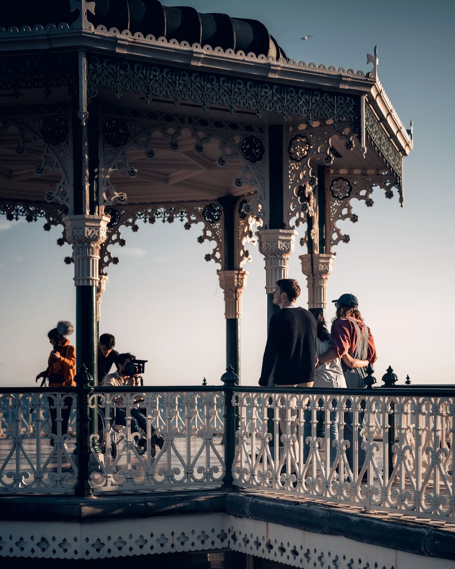 People on a Victorian-style gazebo by the water during sunset, some taking photos and others talking, with clear skies and a bird flying in the background.