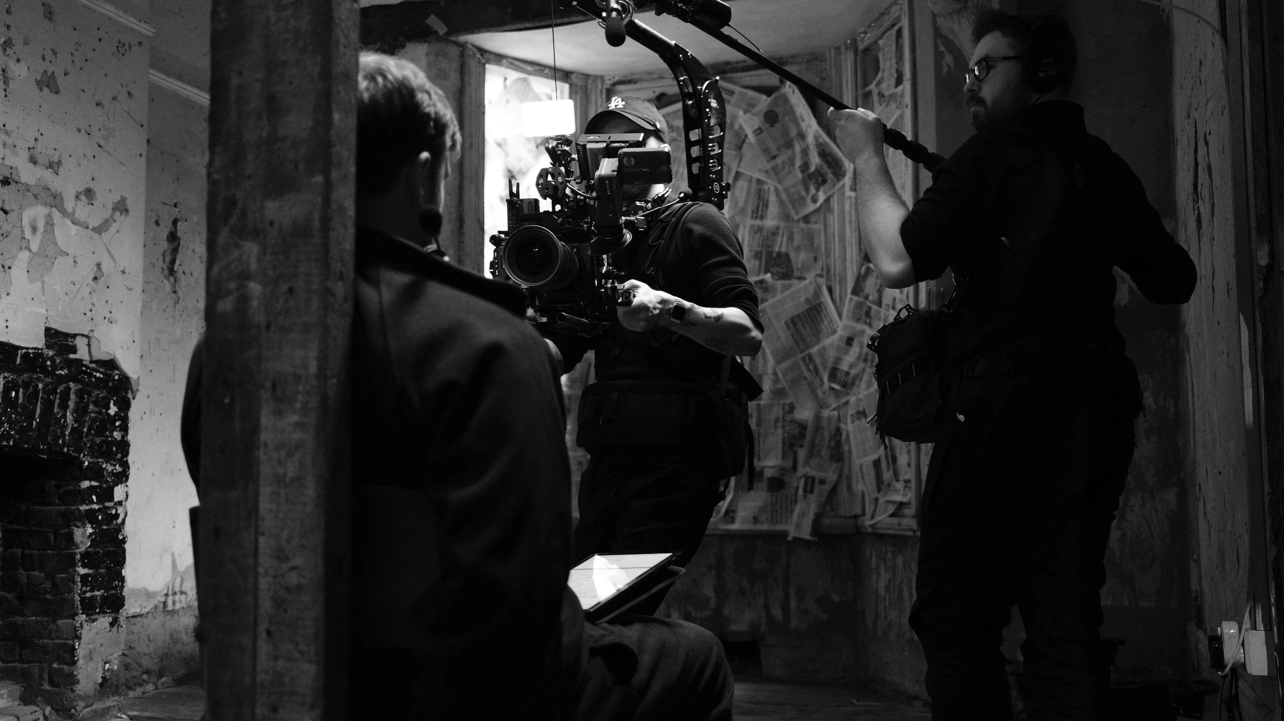 Behind-the-scenes shot of a film crew filming in an abandoned or rundown building, with a cameraman operating a camera rig and another crew member holding a boom microphone, while a person sits nearby with a tablet.
