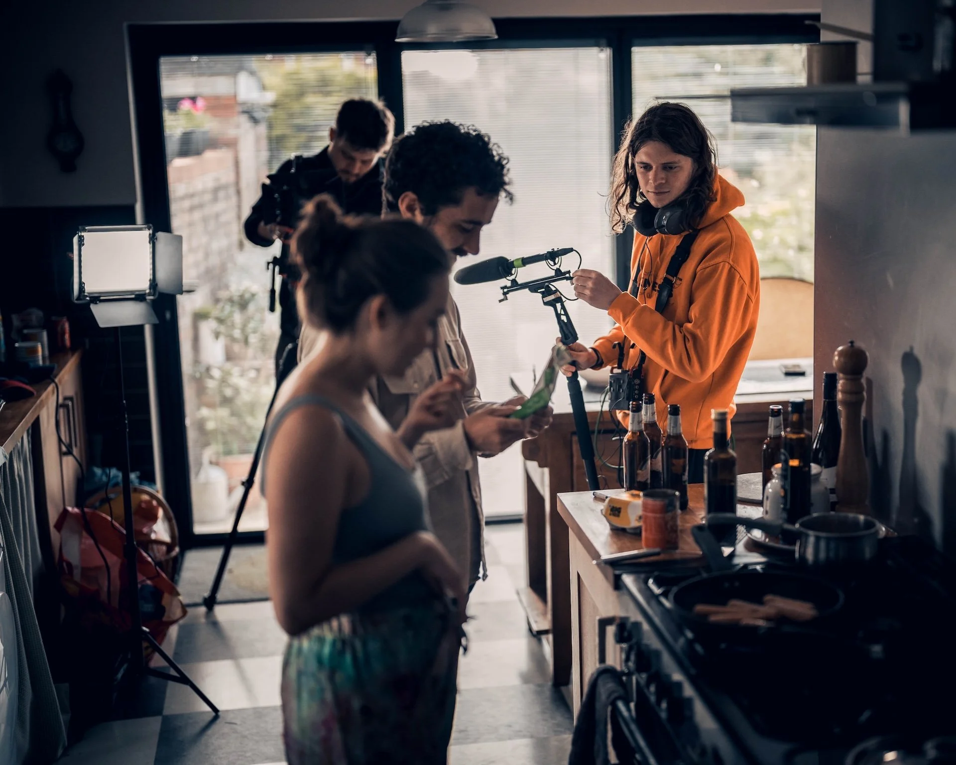Group of four people in a kitchen, preparing drinks, with bottles and bar tools on the counter, and a large window in the background.