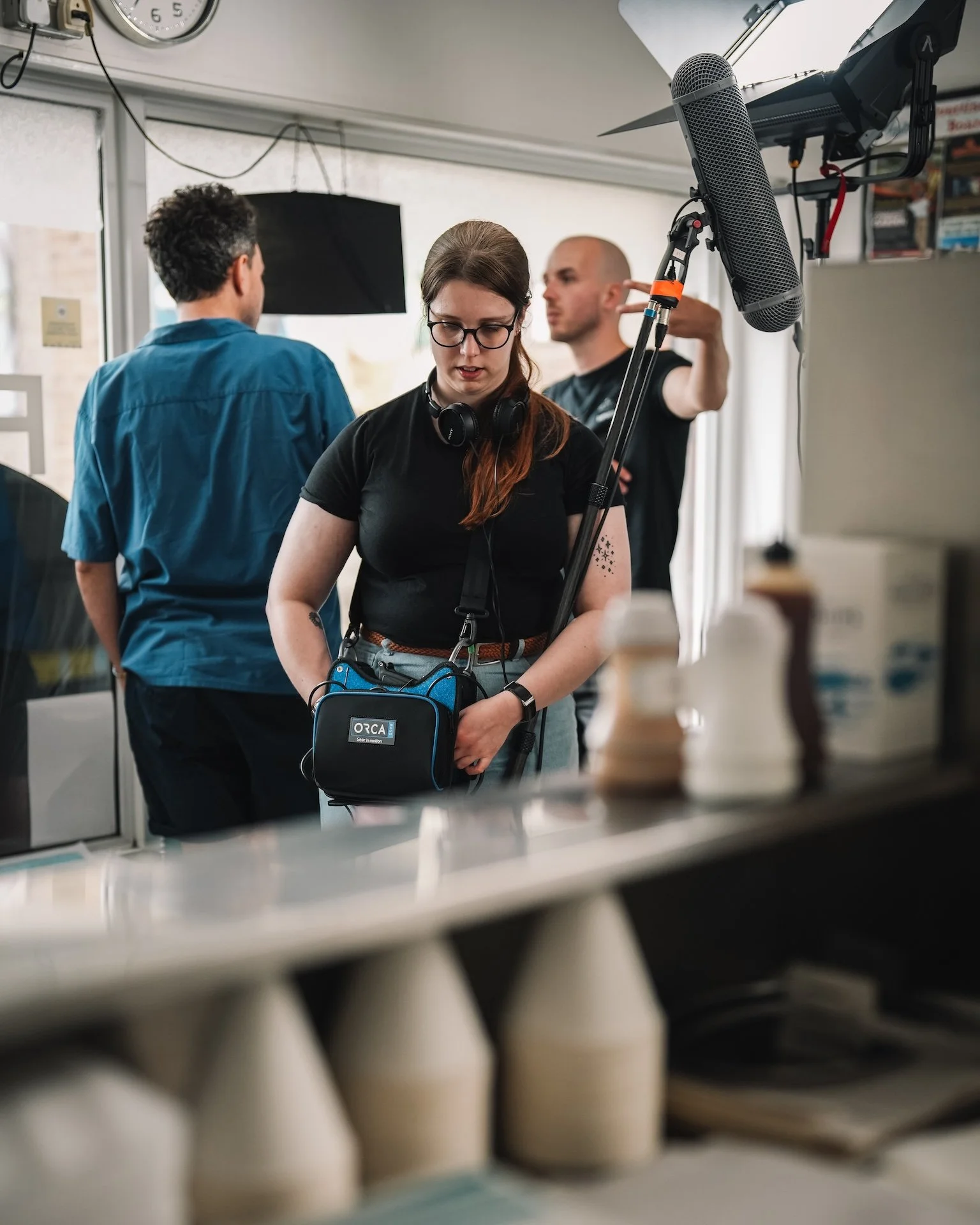 Three people in a kitchen, one woman with glasses and headphones around her neck, two men, one with a shaved head and the other with curly hair, are preparing food or drinks behind a counter. A microphone is hanging from above.