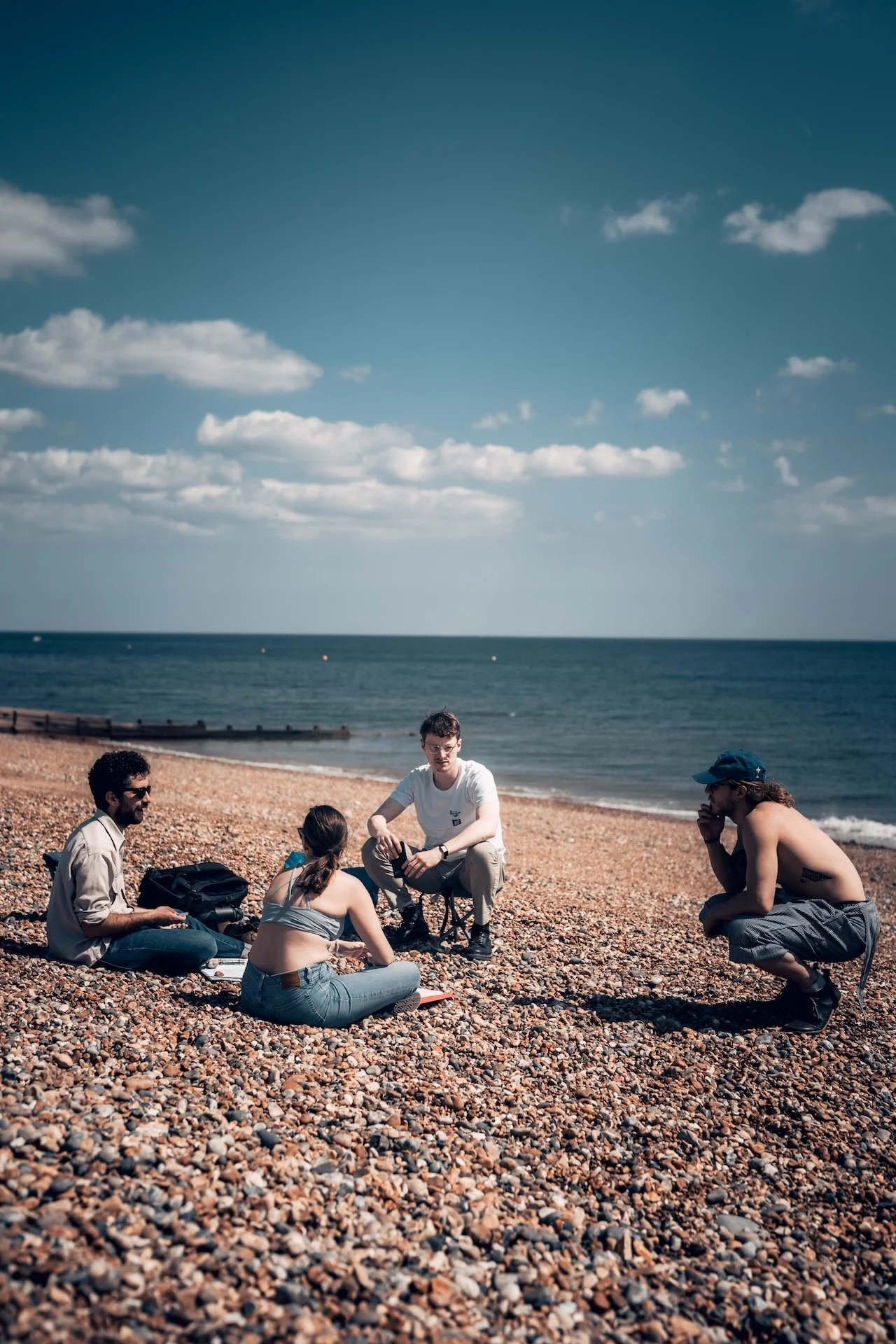 Group of five young adults sitting on a pebble beach having a discussion with the ocean and sky in the background.