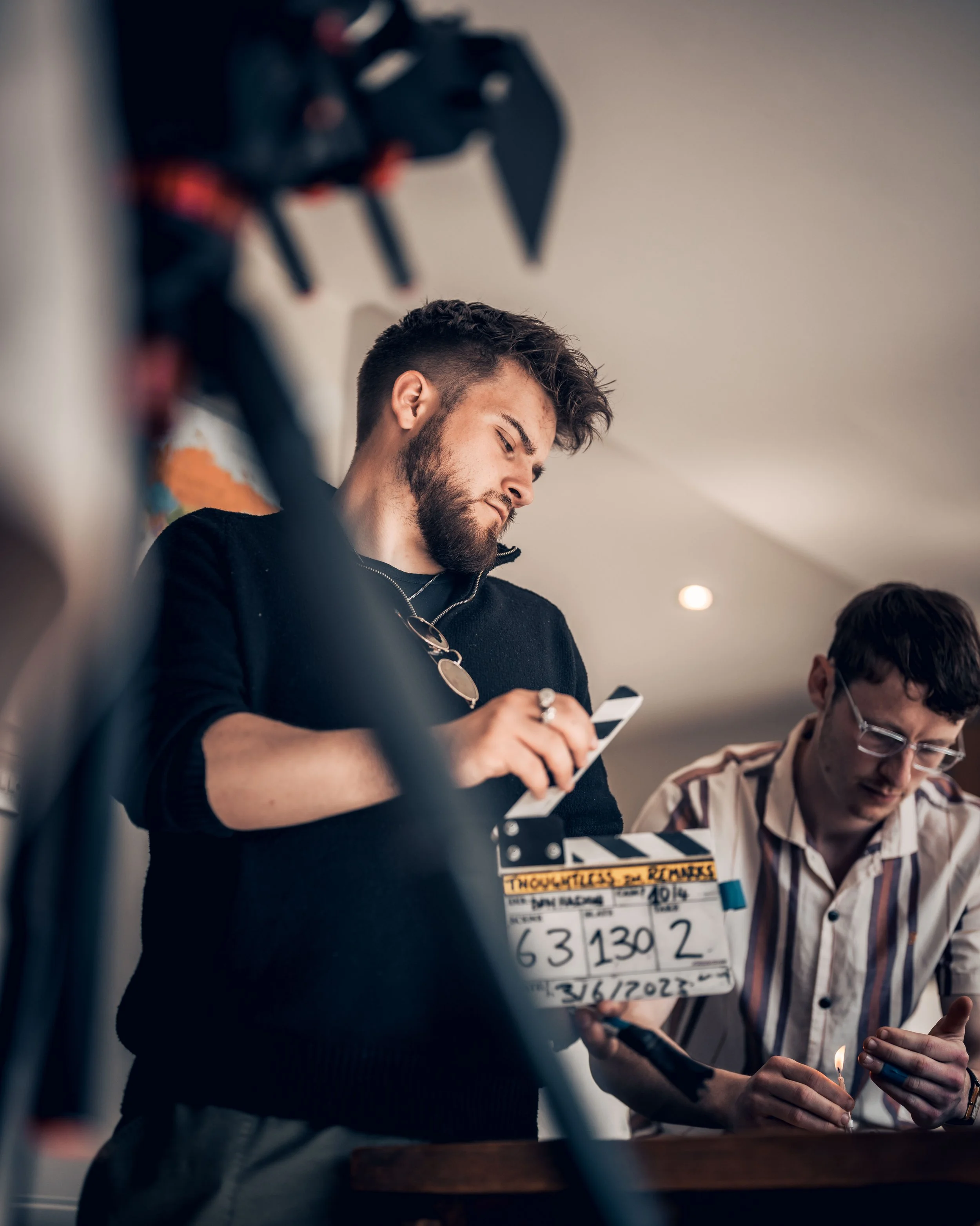 Scene from a film set with a clapperboard showing production details, two men, one holding a lighter and candles, and another using a smartphone.