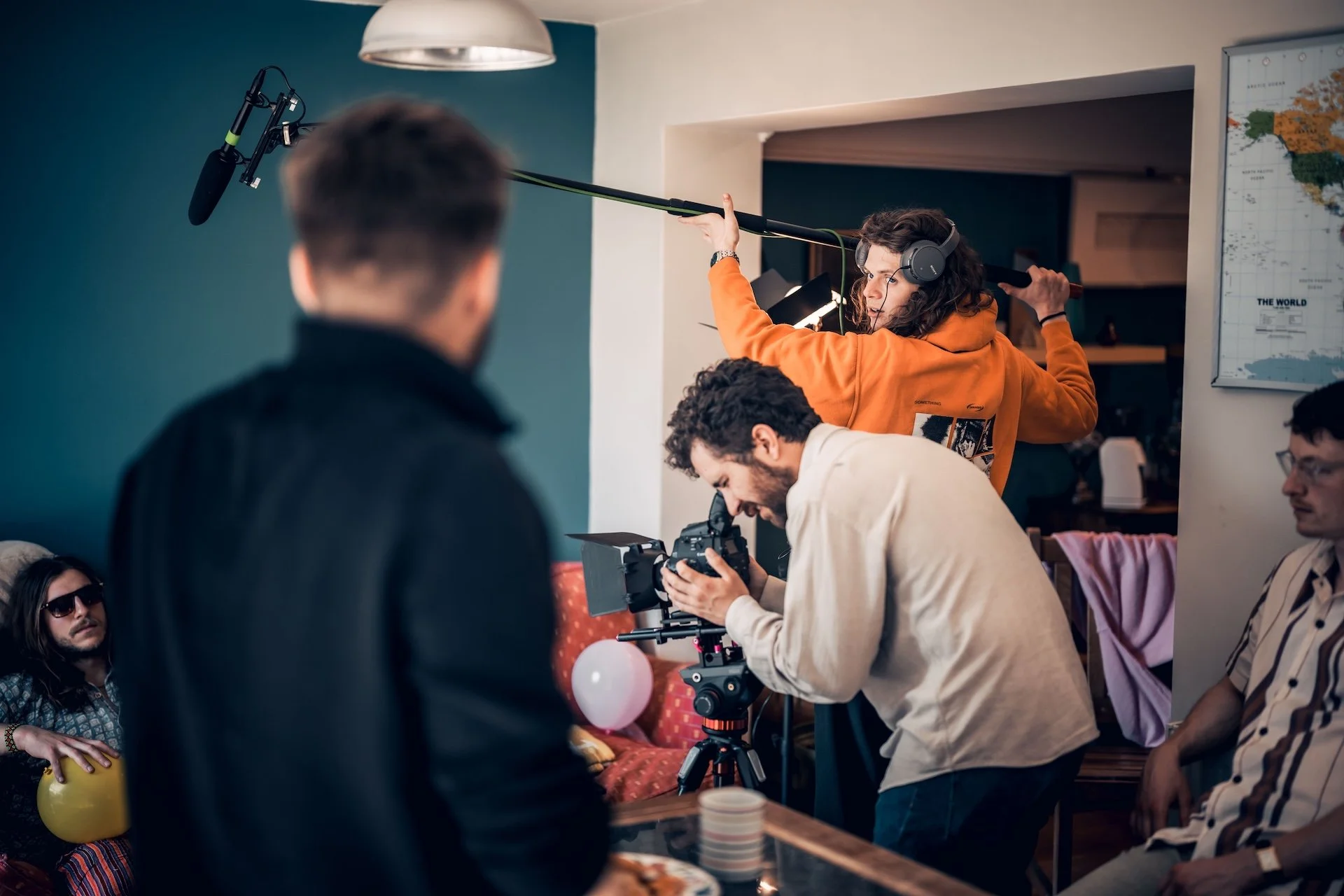 Film crew filming a scene indoors with a camera on tripod, woman in orange hoodie holding boom microphone, crew members working, a woman in sunglasses sitting on a yellow chair, and a man in striped suit sitting nearby.