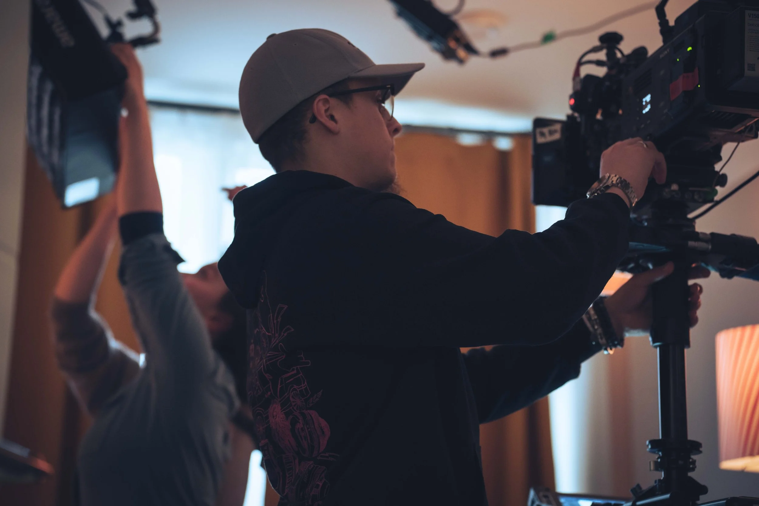 A person operating a professional video camera indoors with another individual partially visible in the foreground.