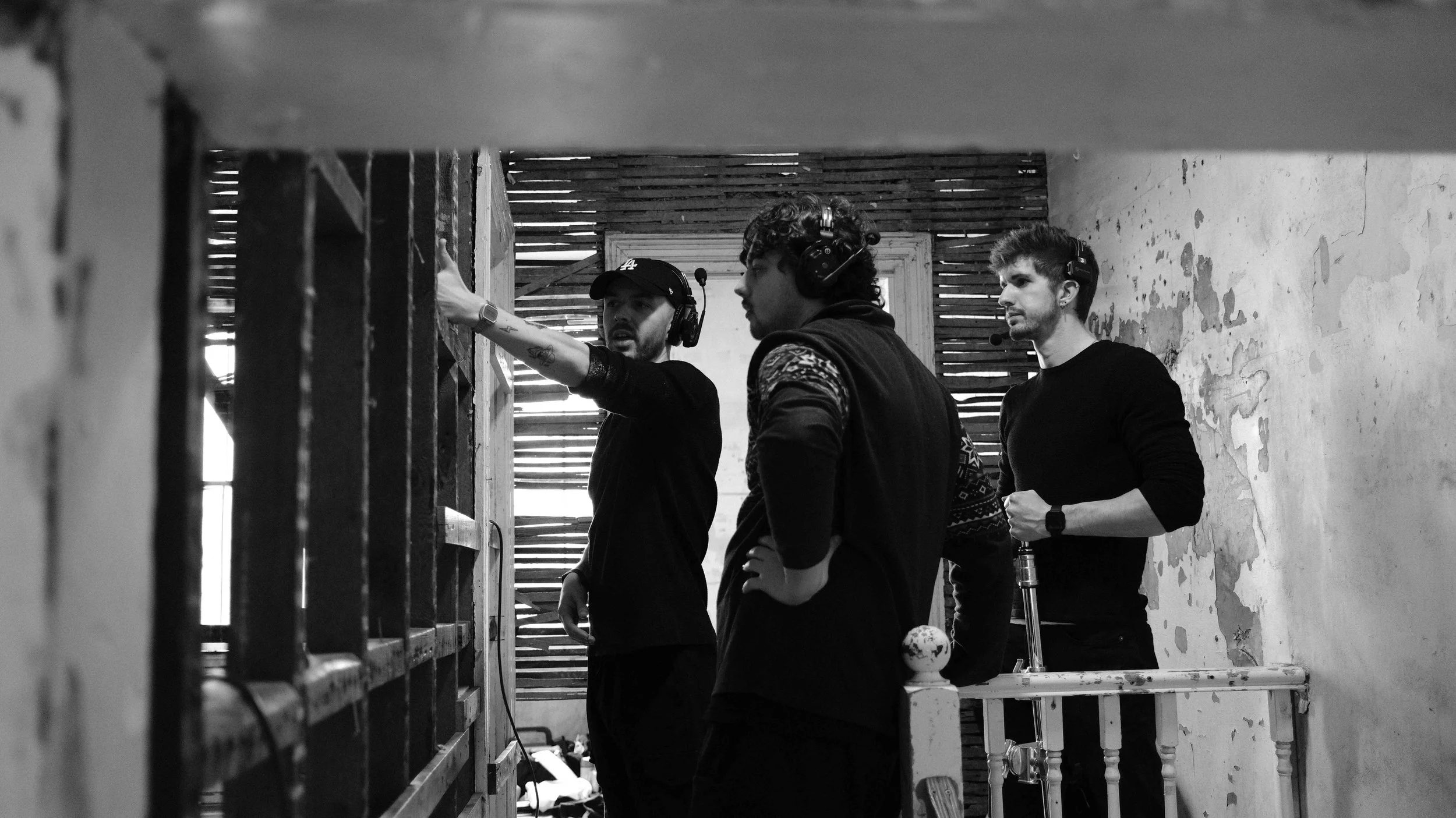 Three men wearing headsets and casual clothing standing in a rustic, partially wooden room, discussing a production or filming setup.